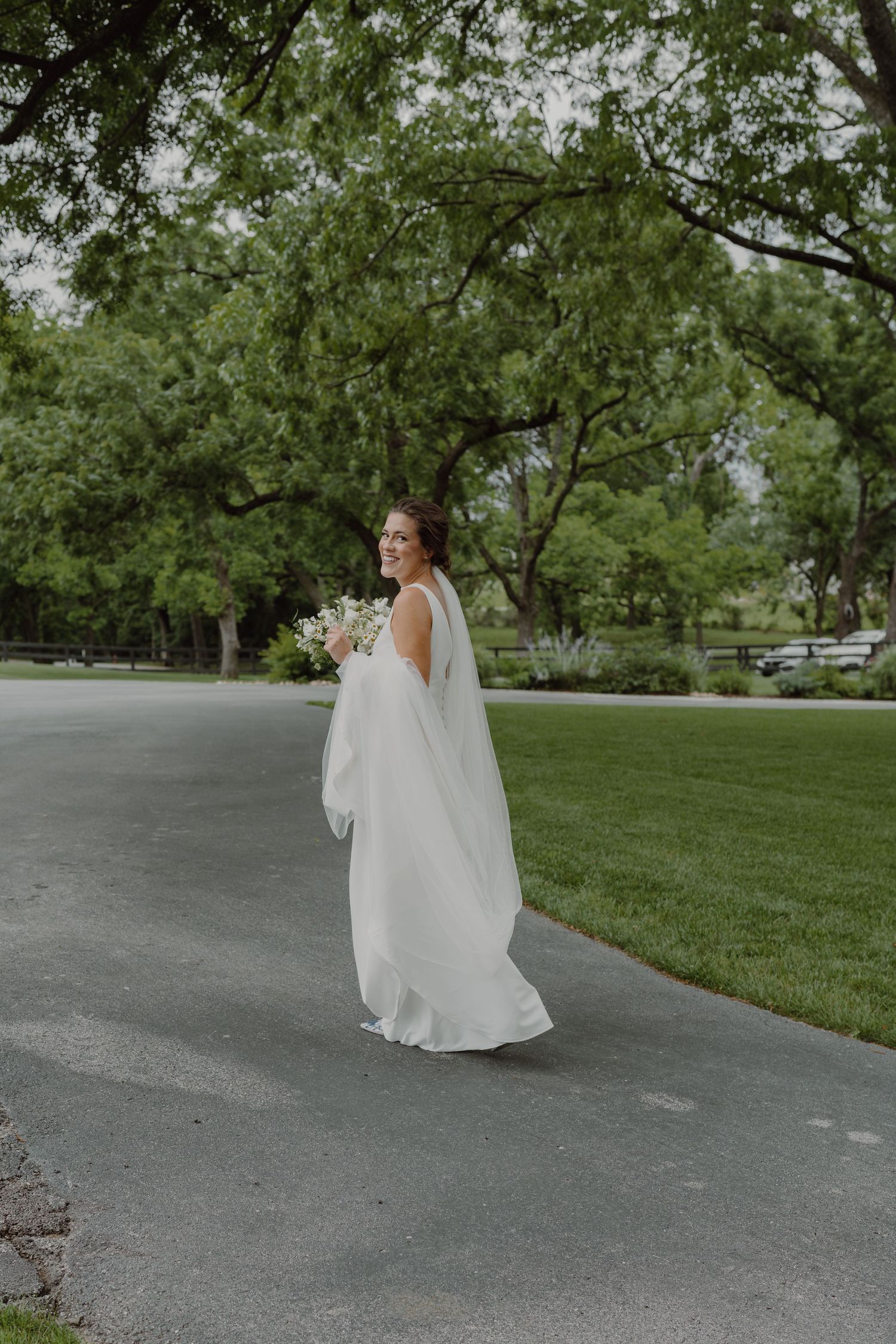 Bride in flowing white dress poses gracefully on concrete driveway surrounded by green trees.
