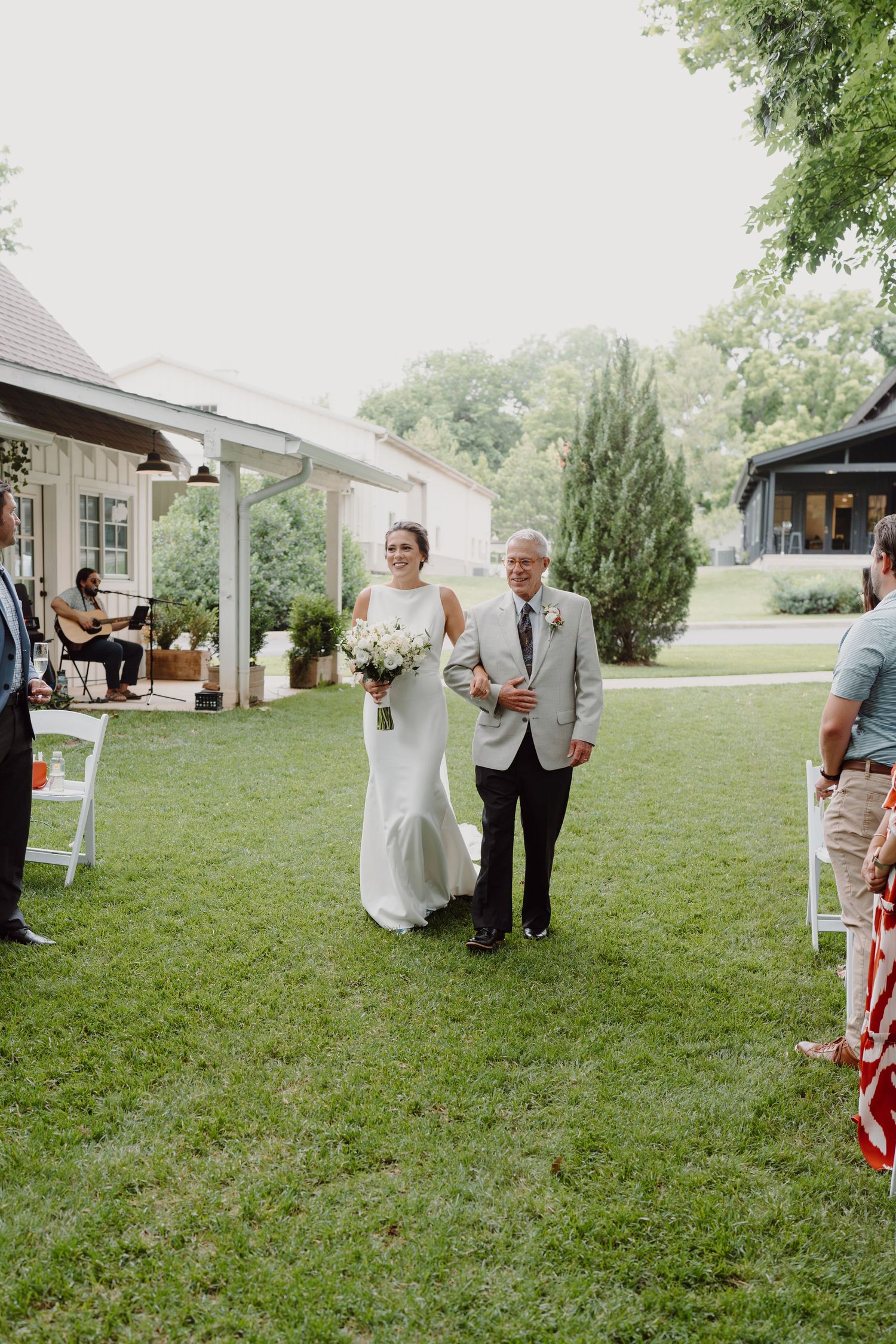 A wedding ceremony processional takes place on a green lawn with white chairs and buildings in background.
