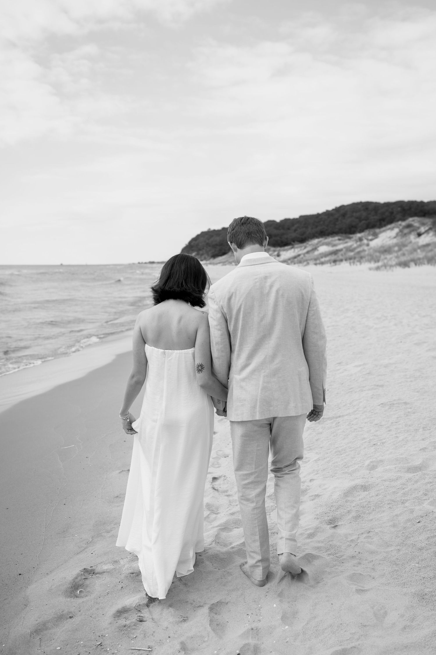 A couple in light colored clothing walks together on a sandy beach in monochrome.