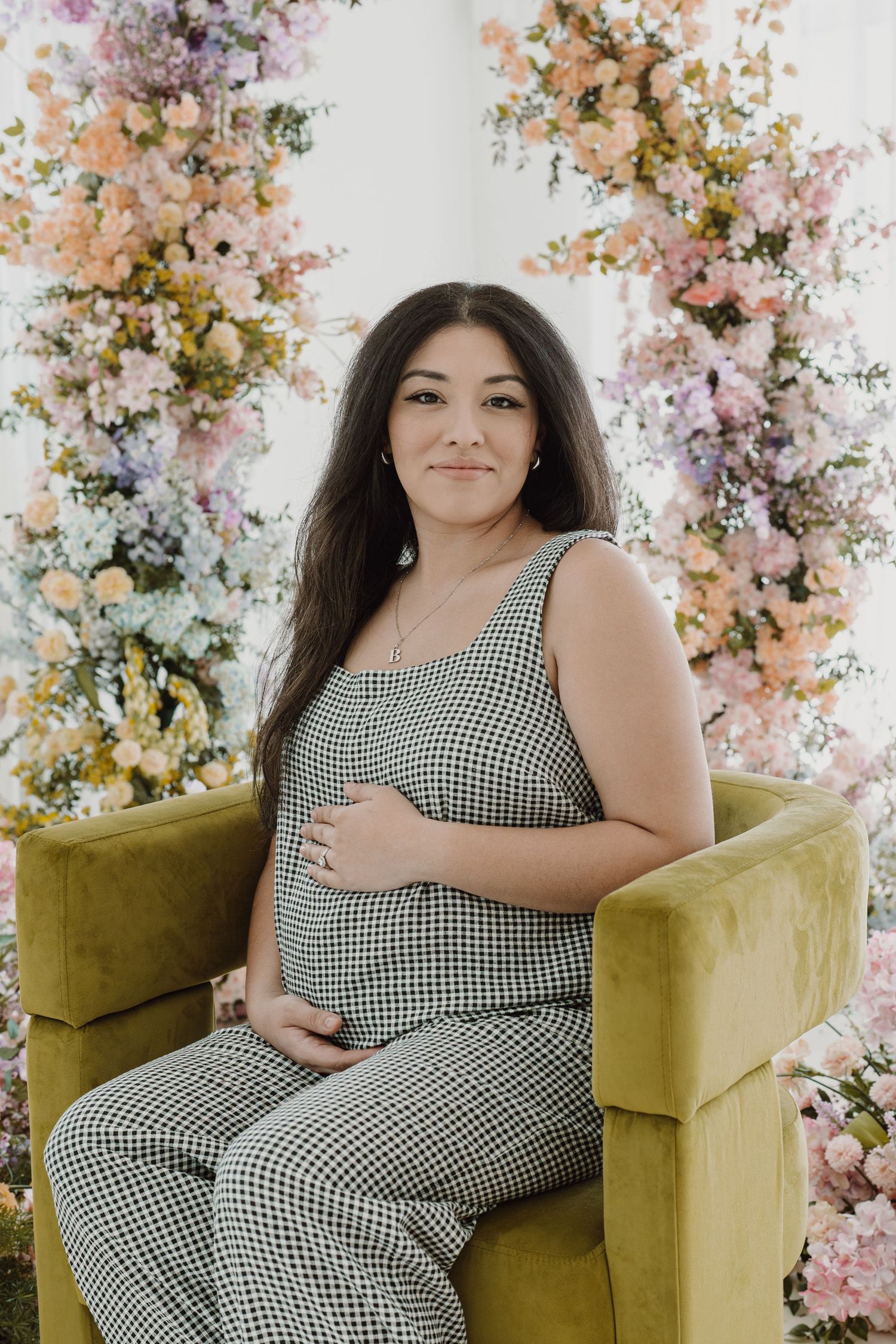 Maternity portrait with woman in checkered jumpsuit seated against floral arch backdrop.