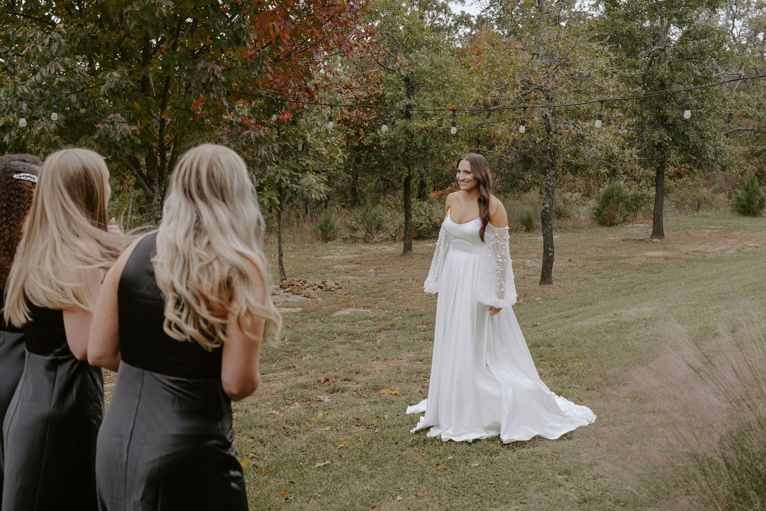 Wedding party in black dresses watches as person in flowing white gown poses in autumn outdoor setting.