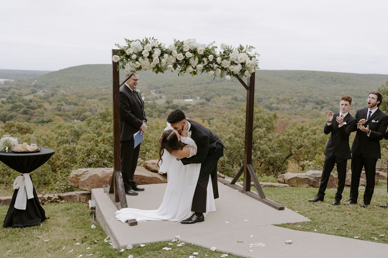 A couple shares their first kiss under a wooden arch decorated with white flowers against a mountain backdrop.