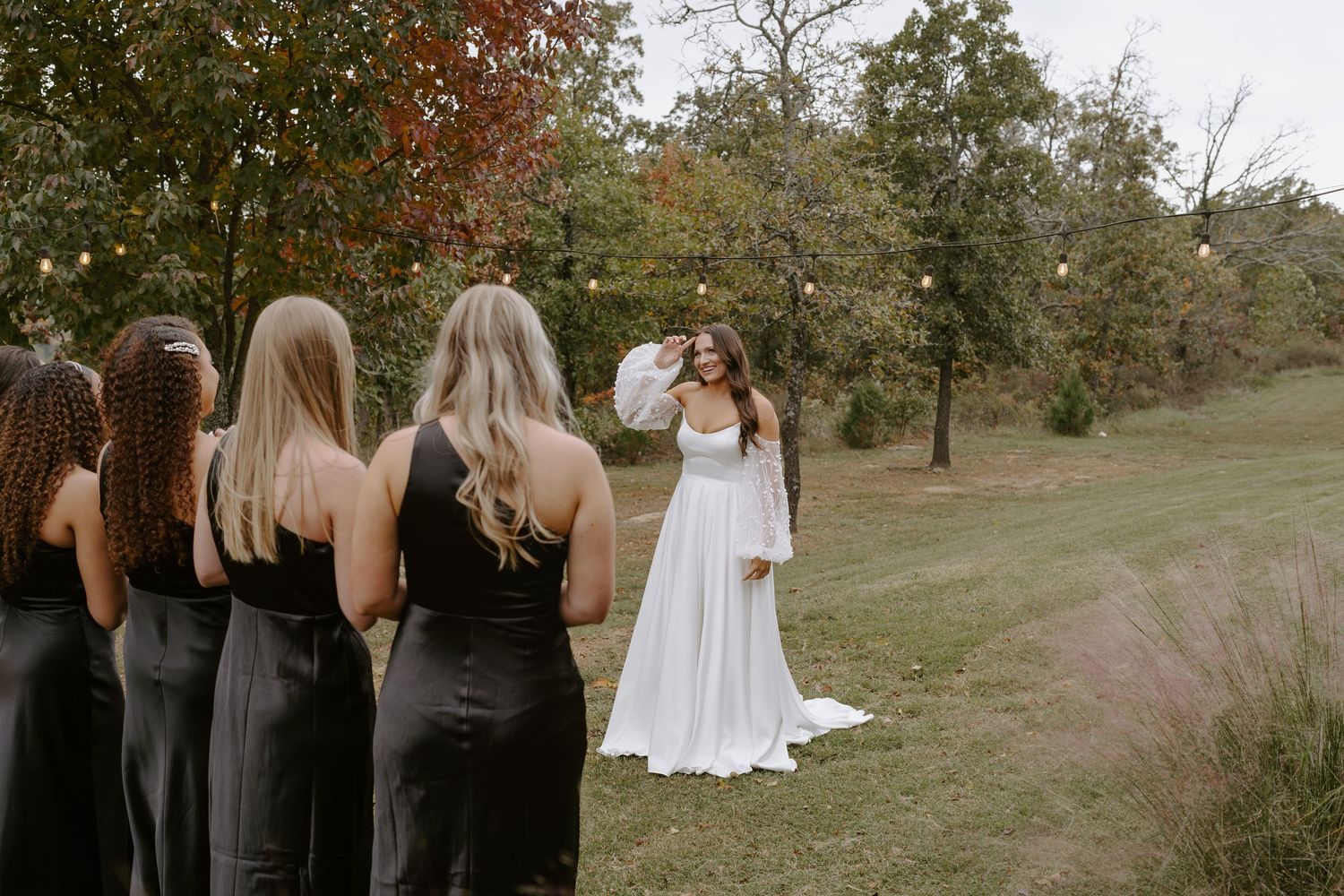 Wedding party in black dresses stands in line watching person in white gown pose on grassy hill.