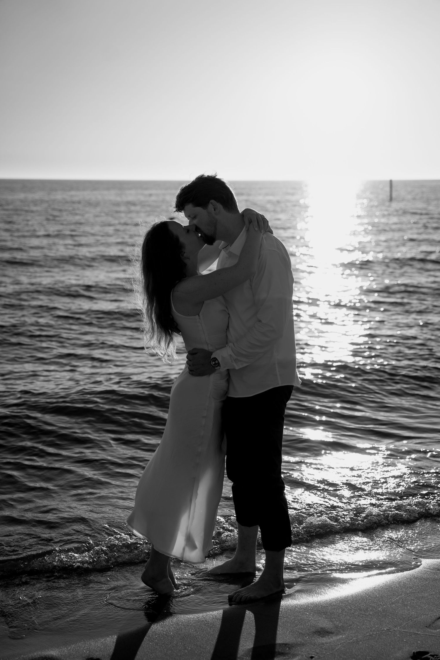 A romantic black and white portrait of a couple embracing on the shoreline.