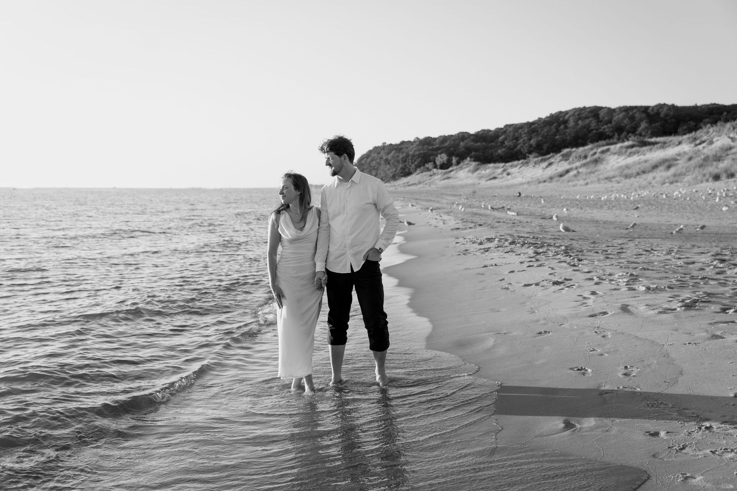 Black and white image of couple strolling together in ankle-deep ocean water.