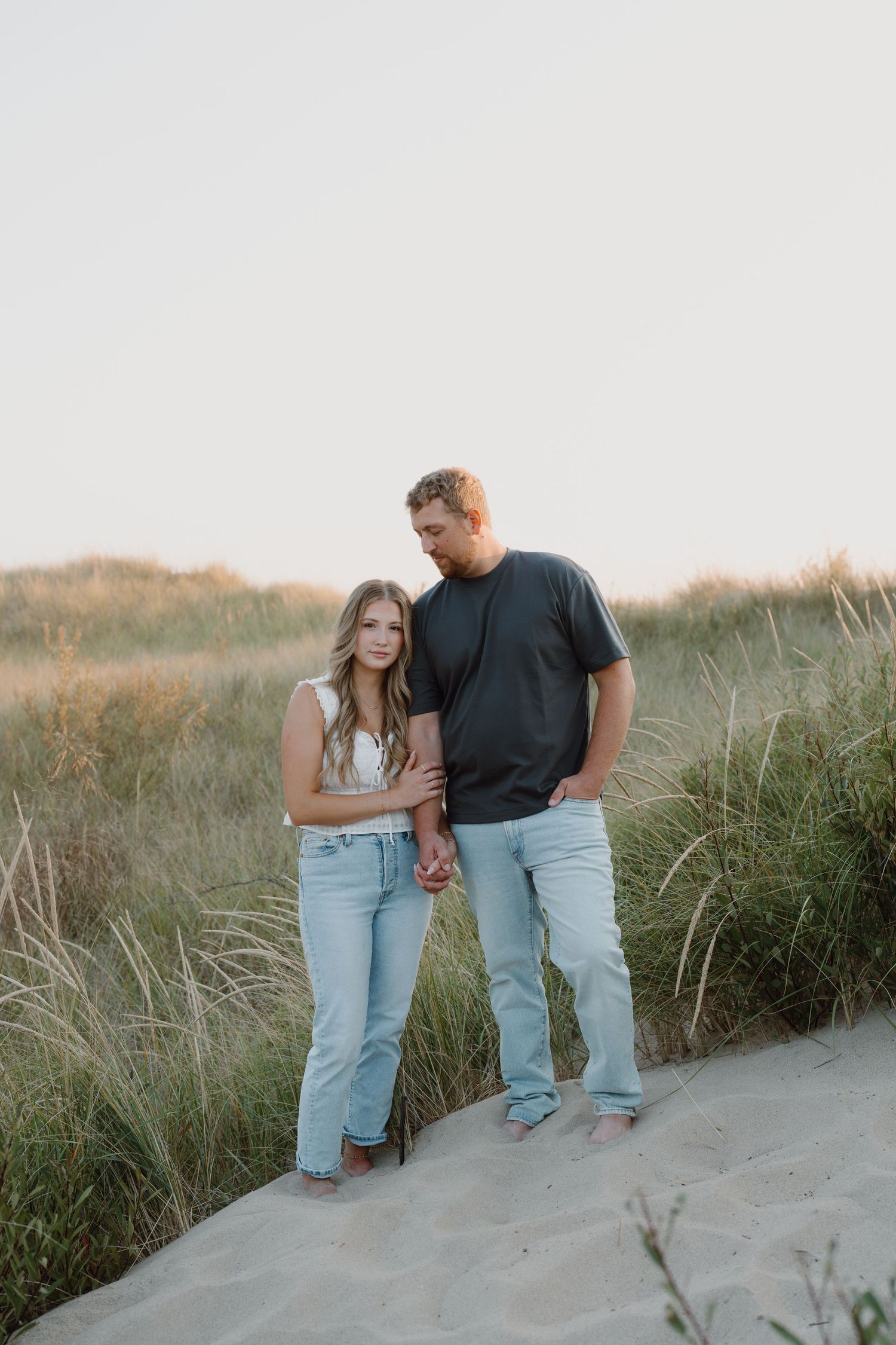 A couple embraces while standing on coastal sand dunes with beach grass in the background.