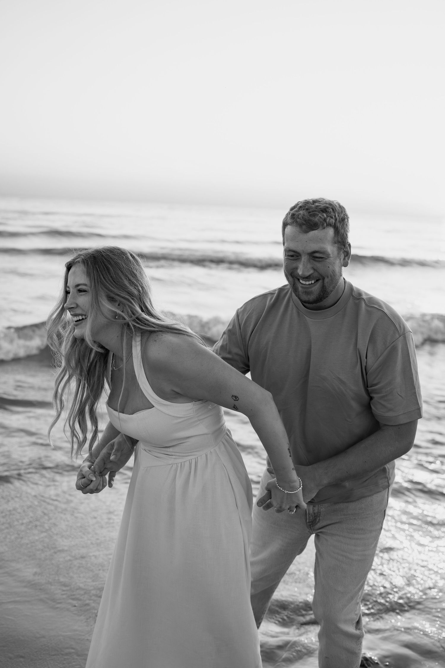 A joyful couple laughs together while walking along the shoreline in black and white.