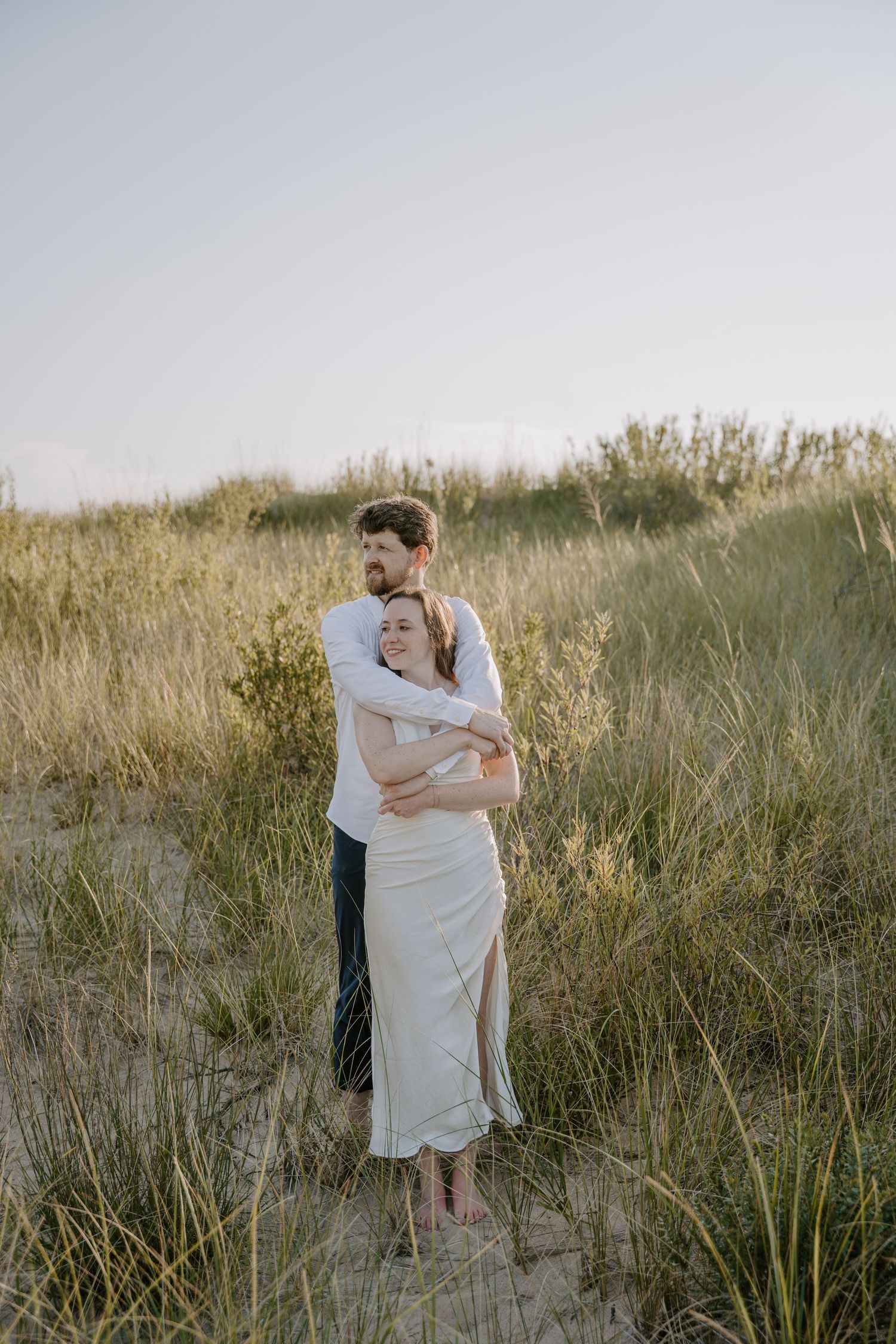 Two people share an intimate moment wrapped in white fabric on a sandy hillside.