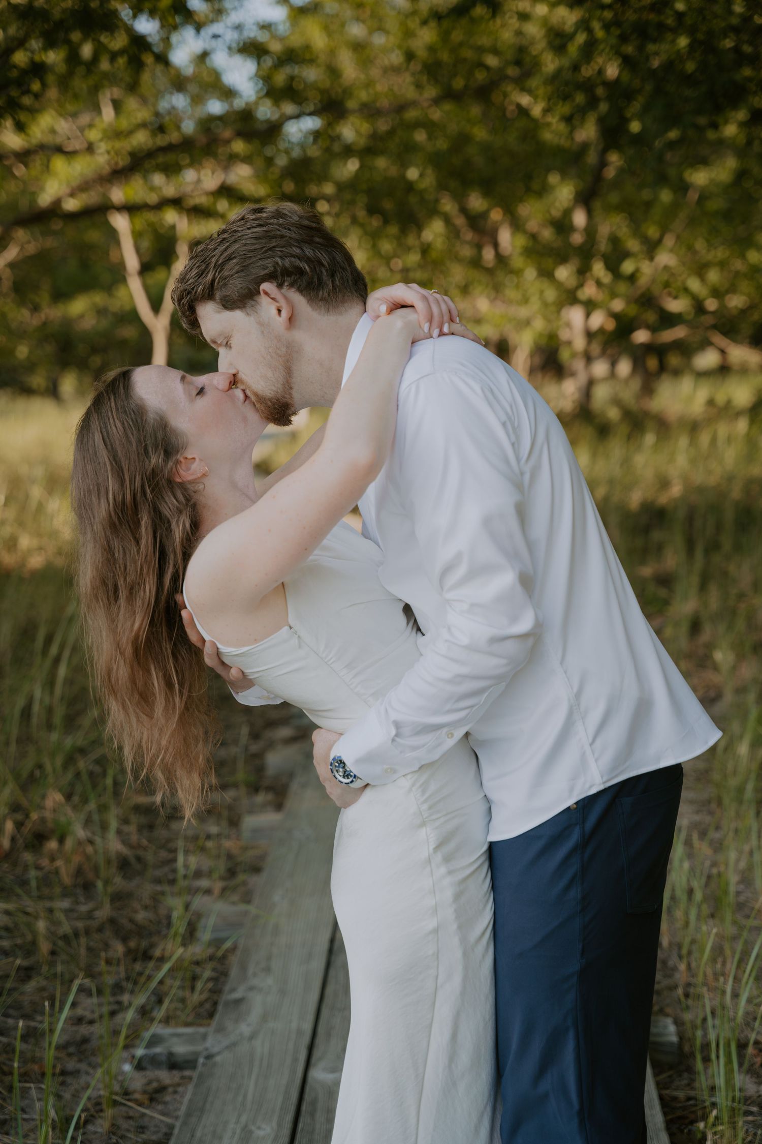 Couple in white and navy formal wear sharing romantic moments among trees and grass.