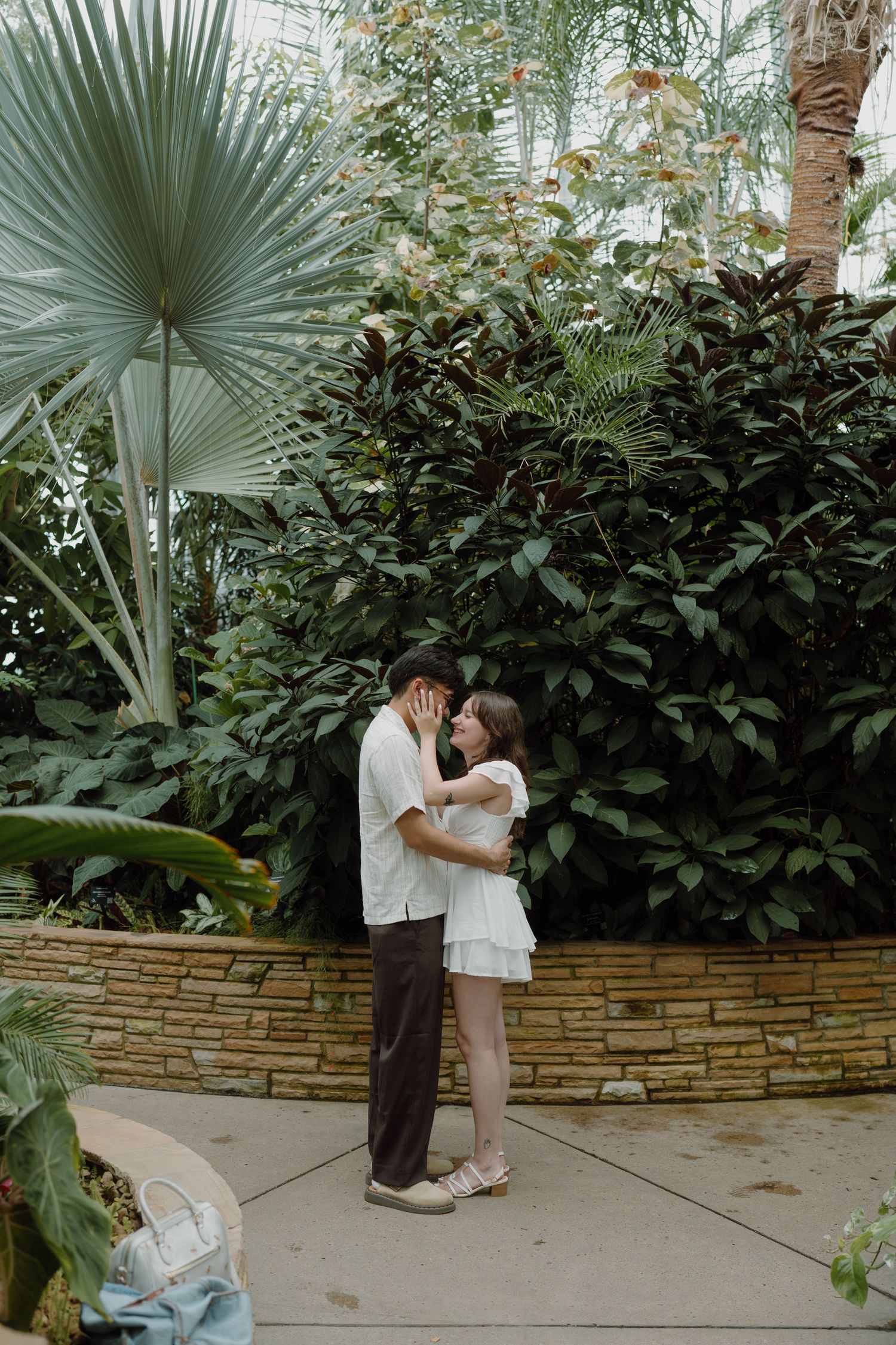A couple shares romantic moments in a garden with palm fronds and stone walls.