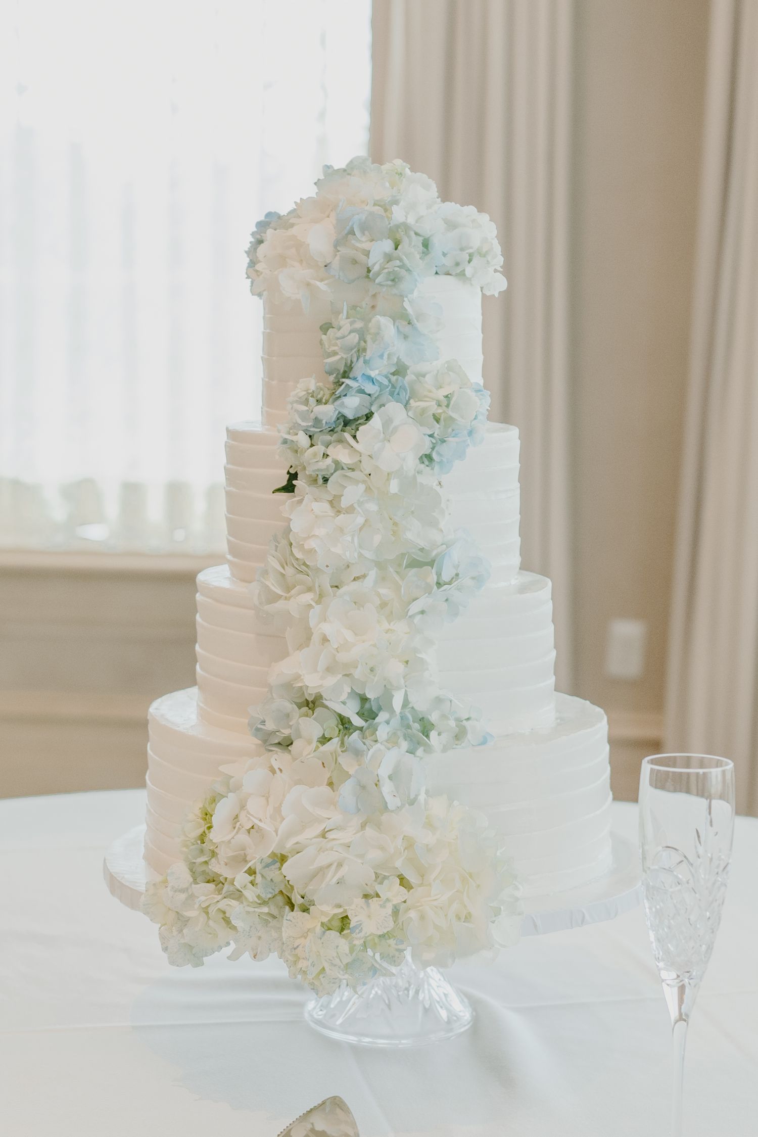 Elegant white tiered wedding cake decorated with delicate hydrangeas and roses on a classic crystal cake stand.