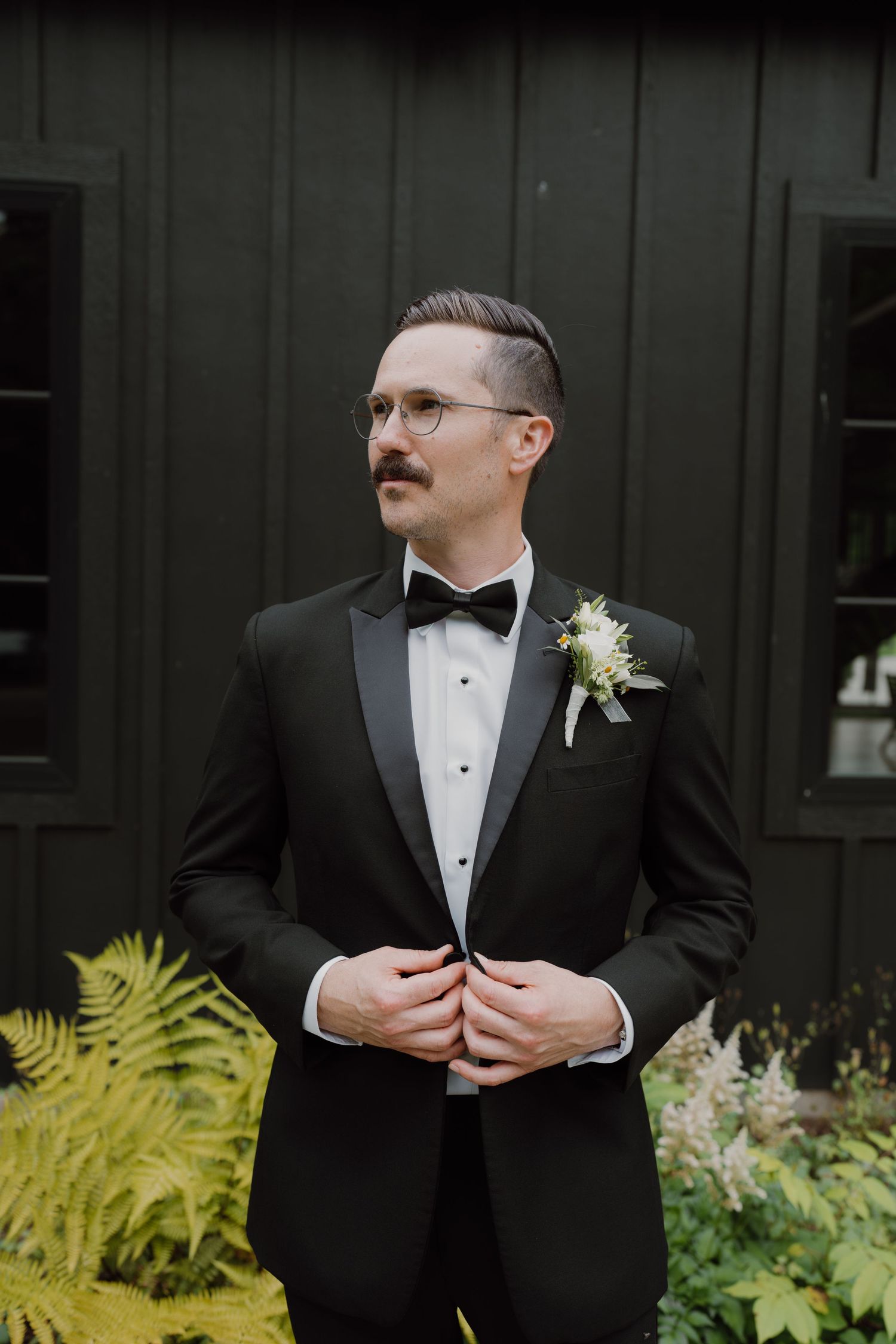 Groom in classic black tuxedo poses against dark wall with yellow ferns in background.