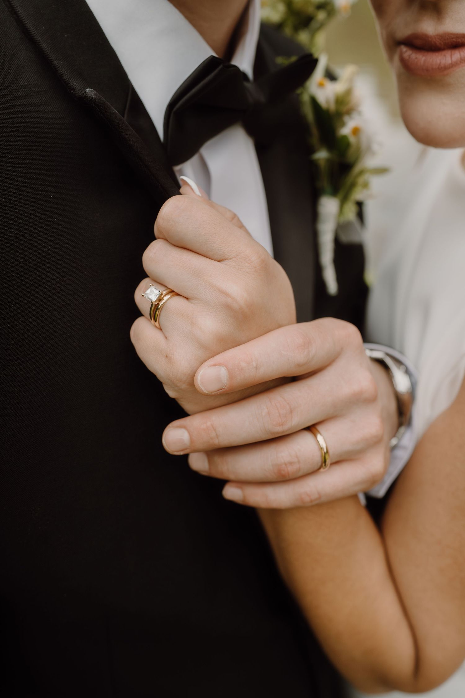 Close-up detail of wedding rings and clasped hands in formal attire.