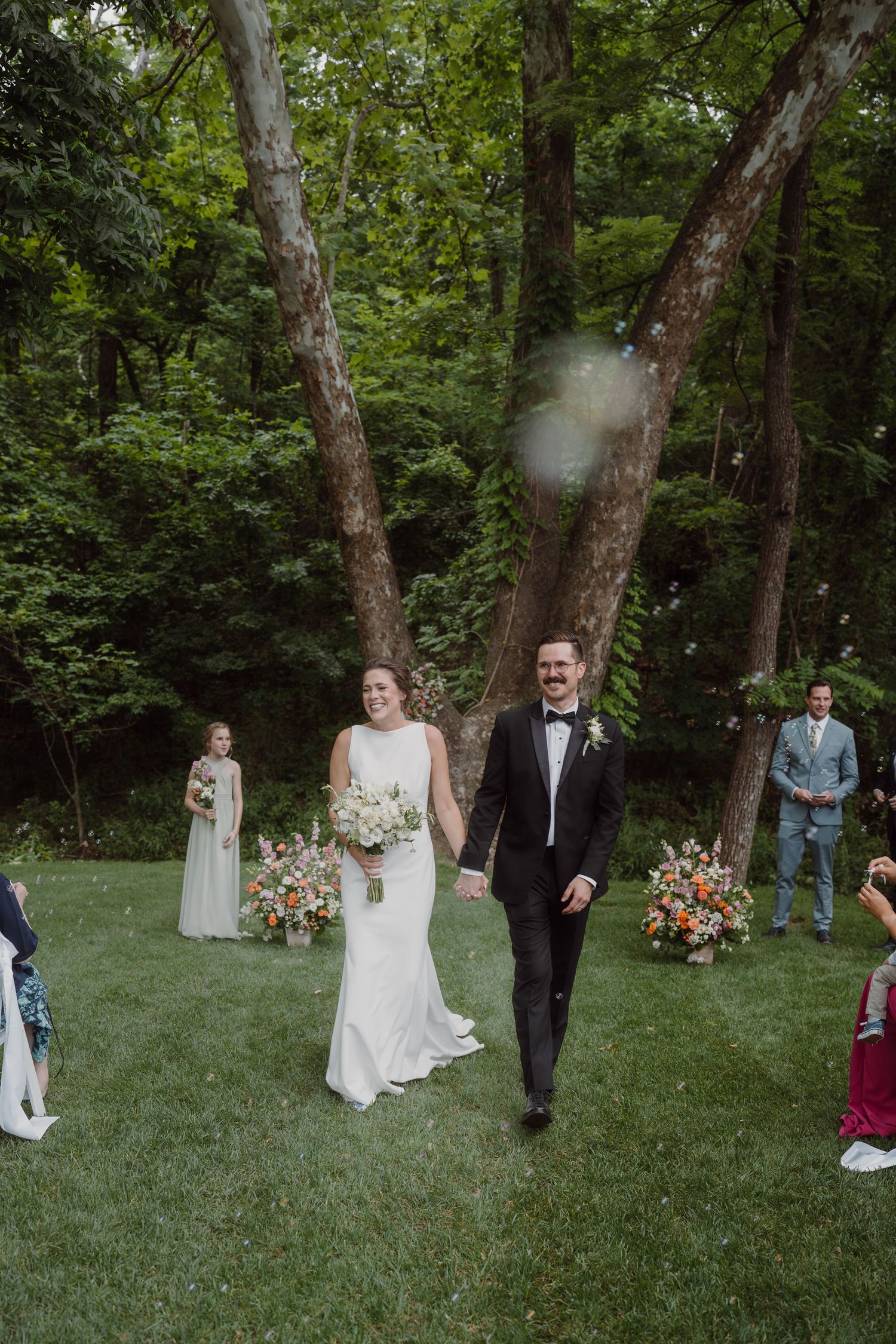 Wedding celebration in a forest clearing with guests seated on white chairs and floral decorations.