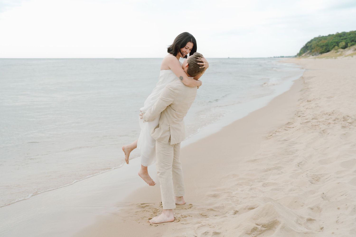 Two people in light colored clothing playfully run and dance along a serene beach landscape.