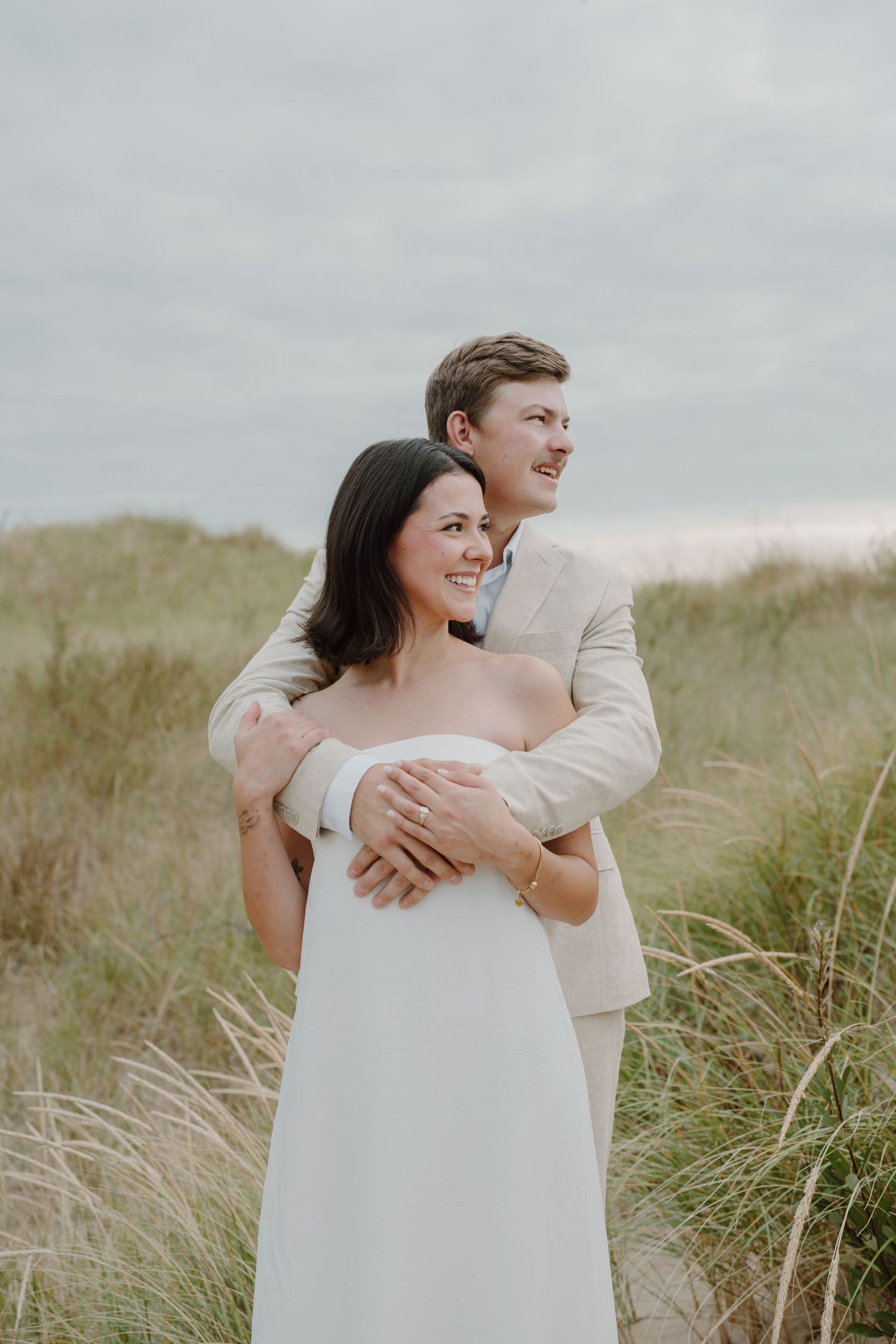 Candid beach wedding portraits showing couple's genuine laughter and love among coastal grasses.