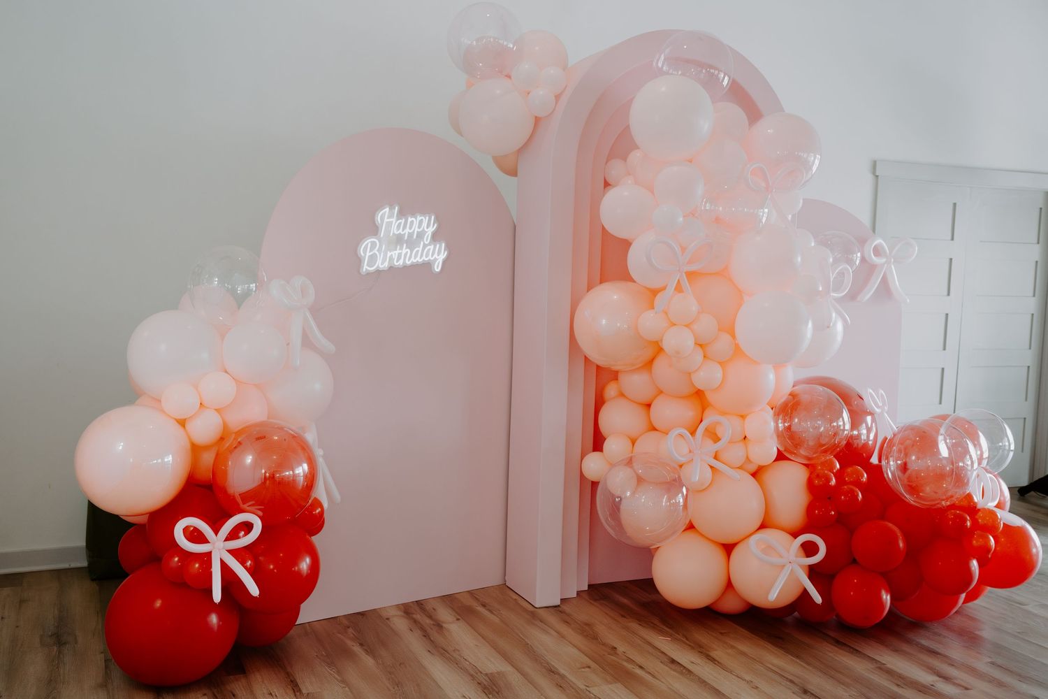 Pink and red balloon arch installation with white bows against a light wall.