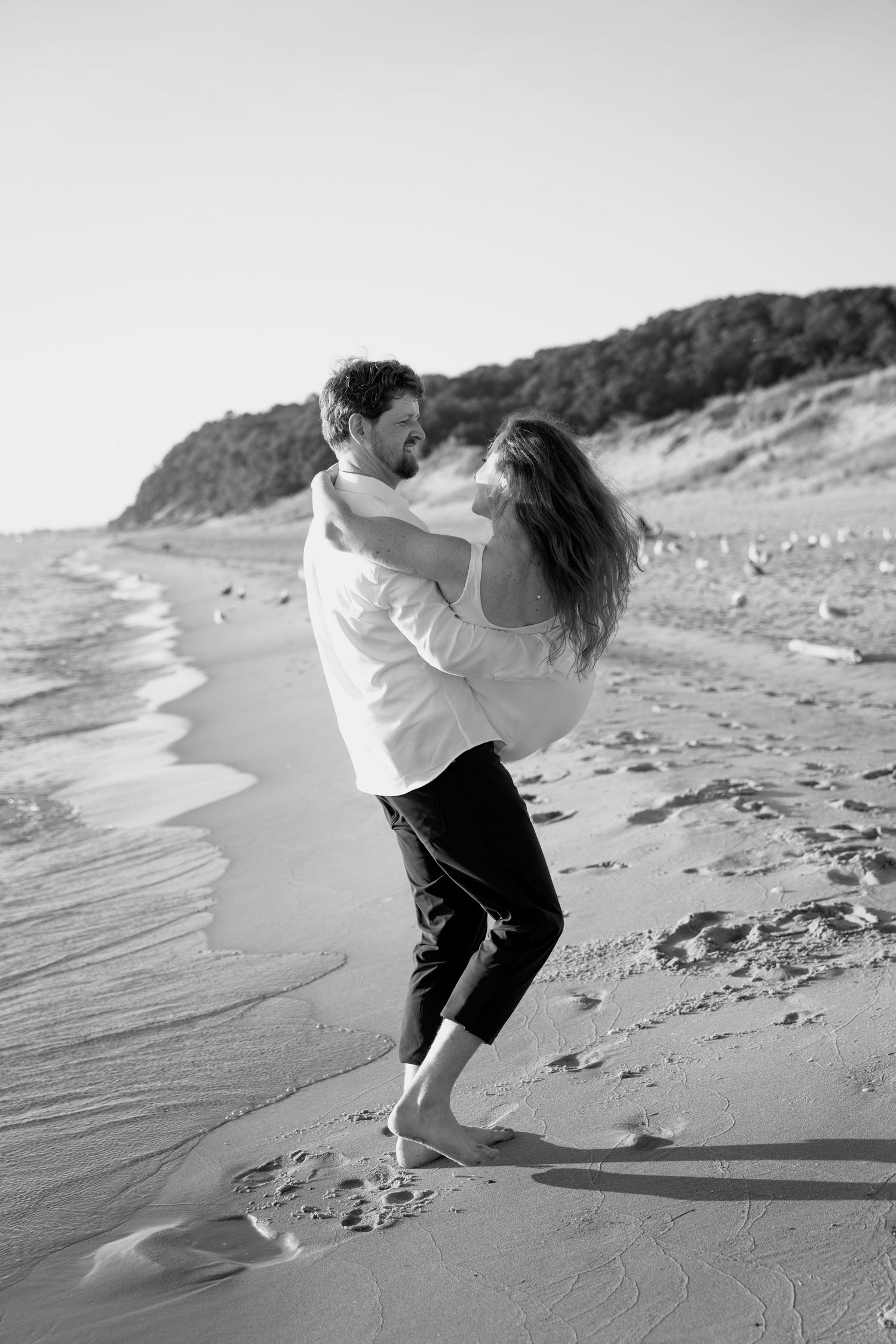 Black and white photo of a playful embrace on a beach with cliffs in the distance.