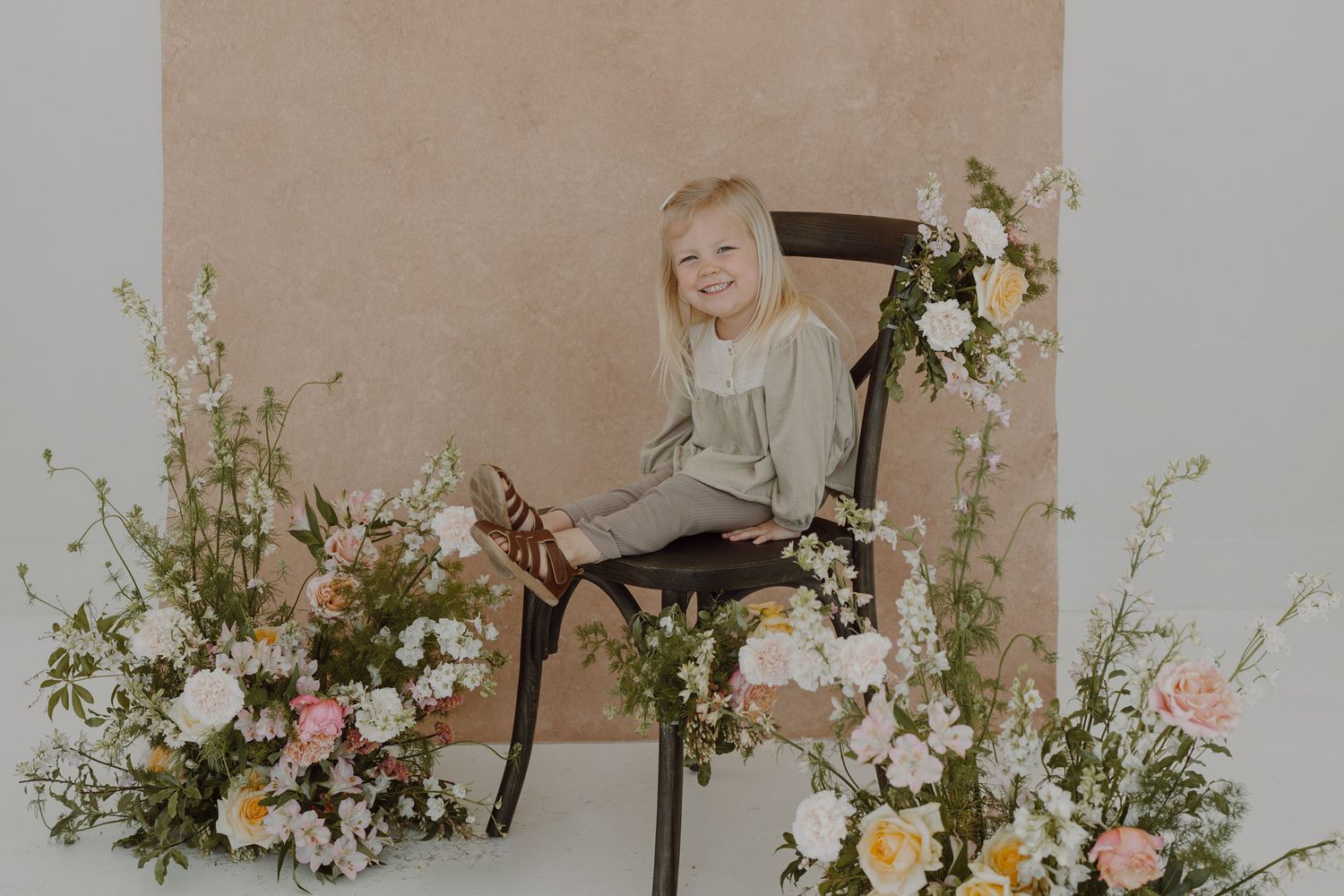 Portrait setup featuring pastel floral arrangements and wooden chair against beige backdrop.