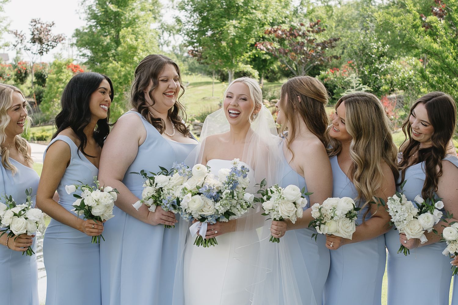 Bride in white gown laughs with bridesmaids in light blue dresses holding white floral bouquets outdoors.