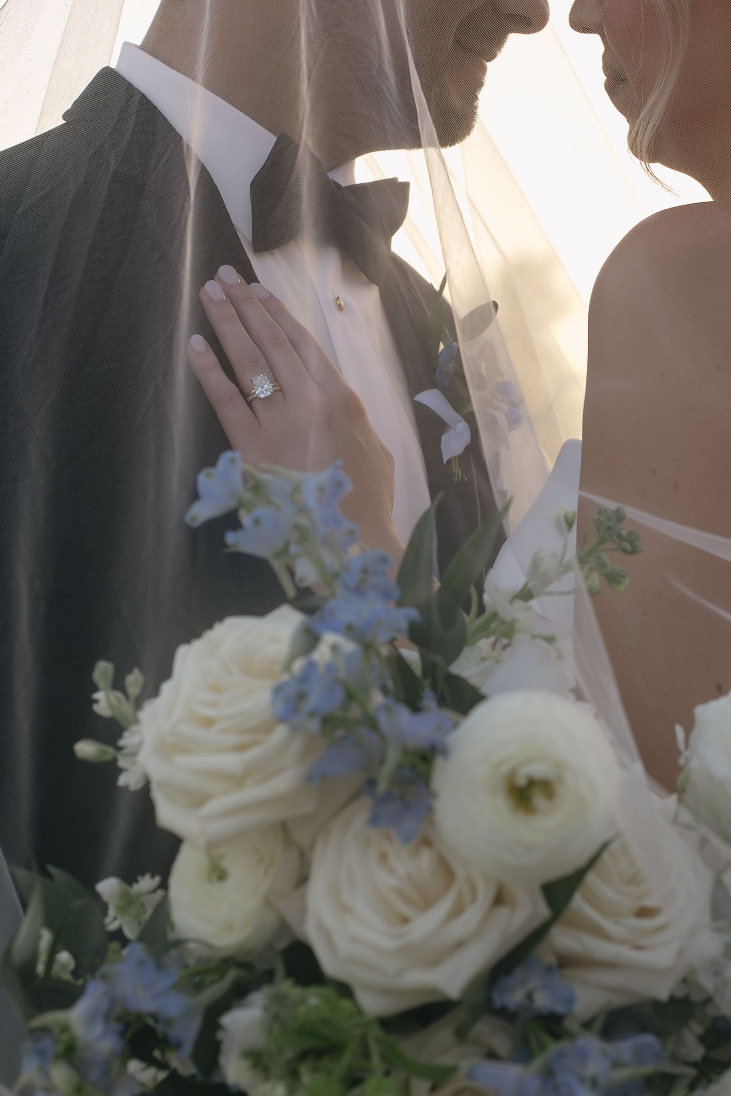Bride and groom under veil with white and blue bouquet, diamond engagement ring visible, romantic wedding portrait.