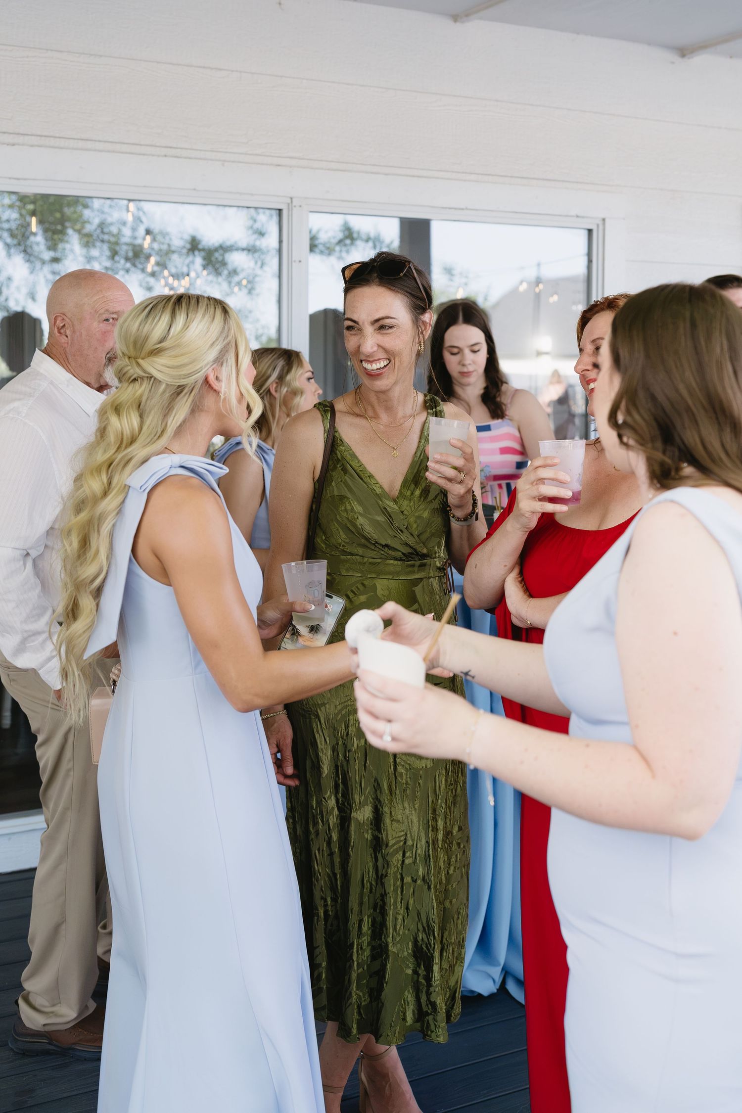 Women in elegant dresses laughing and socializing at a wedding cocktail hour, holding drinks outdoors.