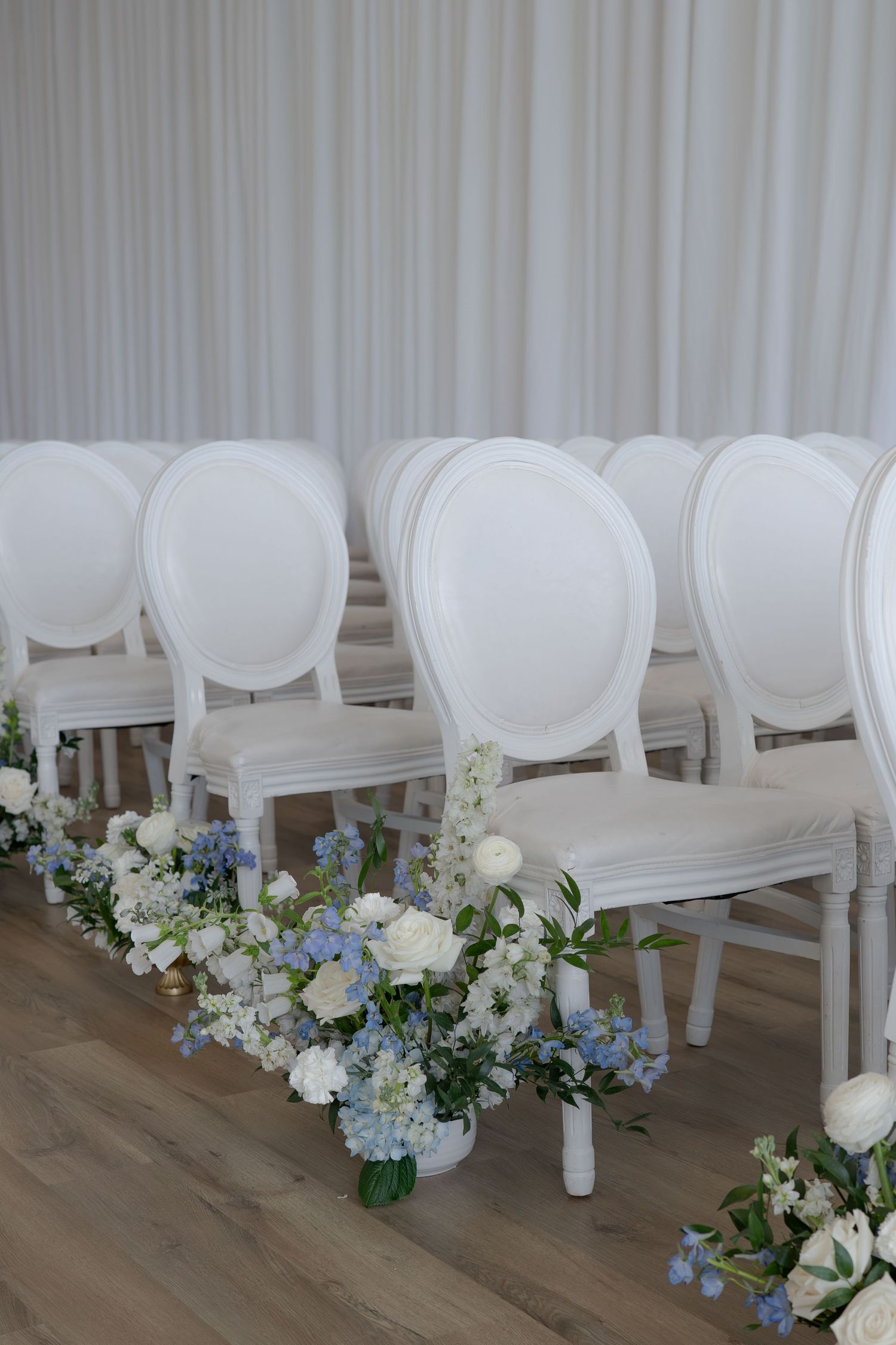 Elegant white Louis XVI chairs lined up with blue and white floral arrangements on the floor for a wedding ceremony.