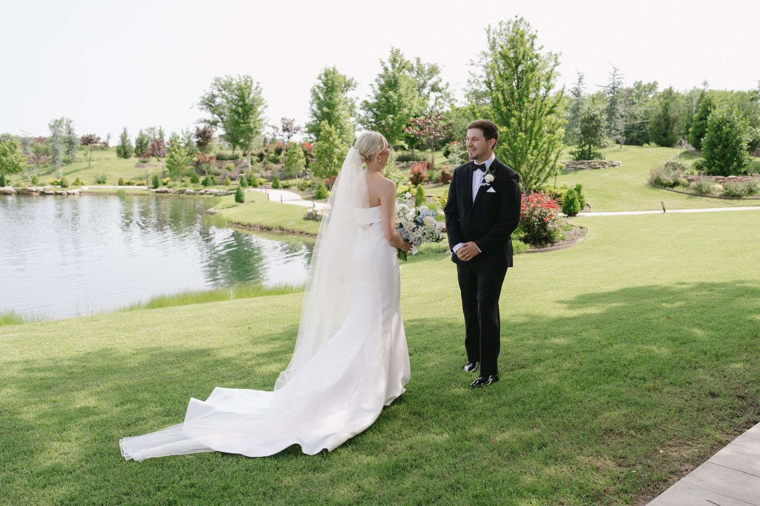 Bride and groom share a first look moment on lush green lawn beside a serene pond at an elegant outdoor wedding venue.
