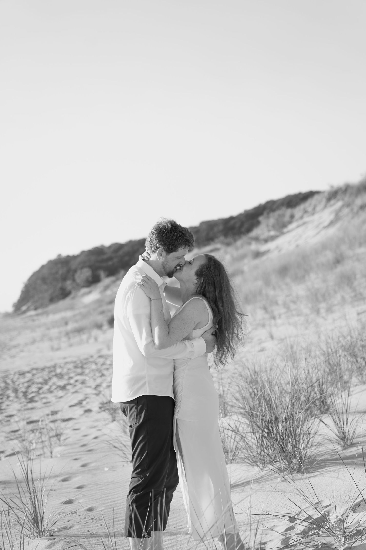A black and white portrait of a couple sharing an intimate moment on a coastal dune.