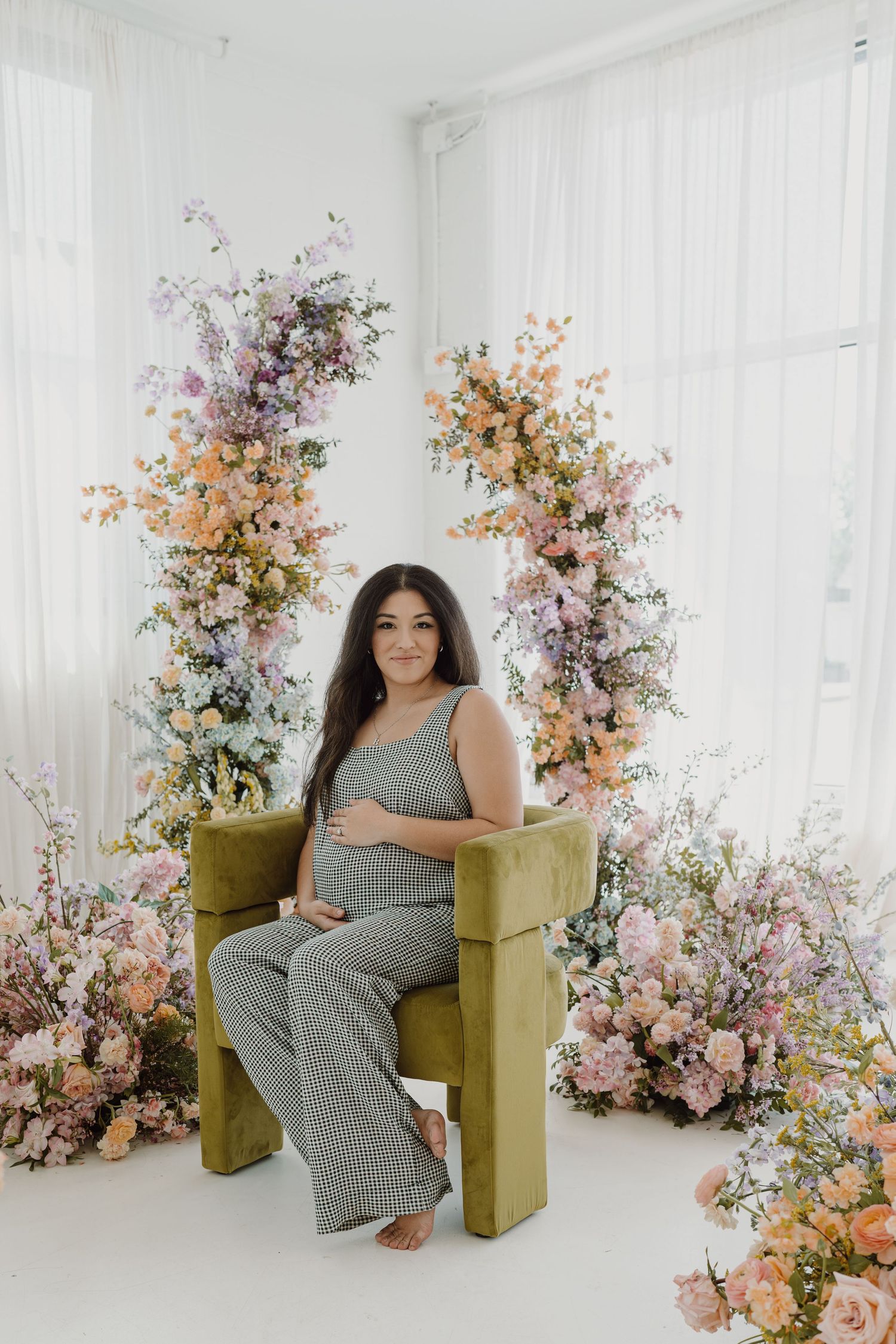 Maternity photoshoot featuring woman in gray jumpsuit seated on mustard chair with floral arrangements.