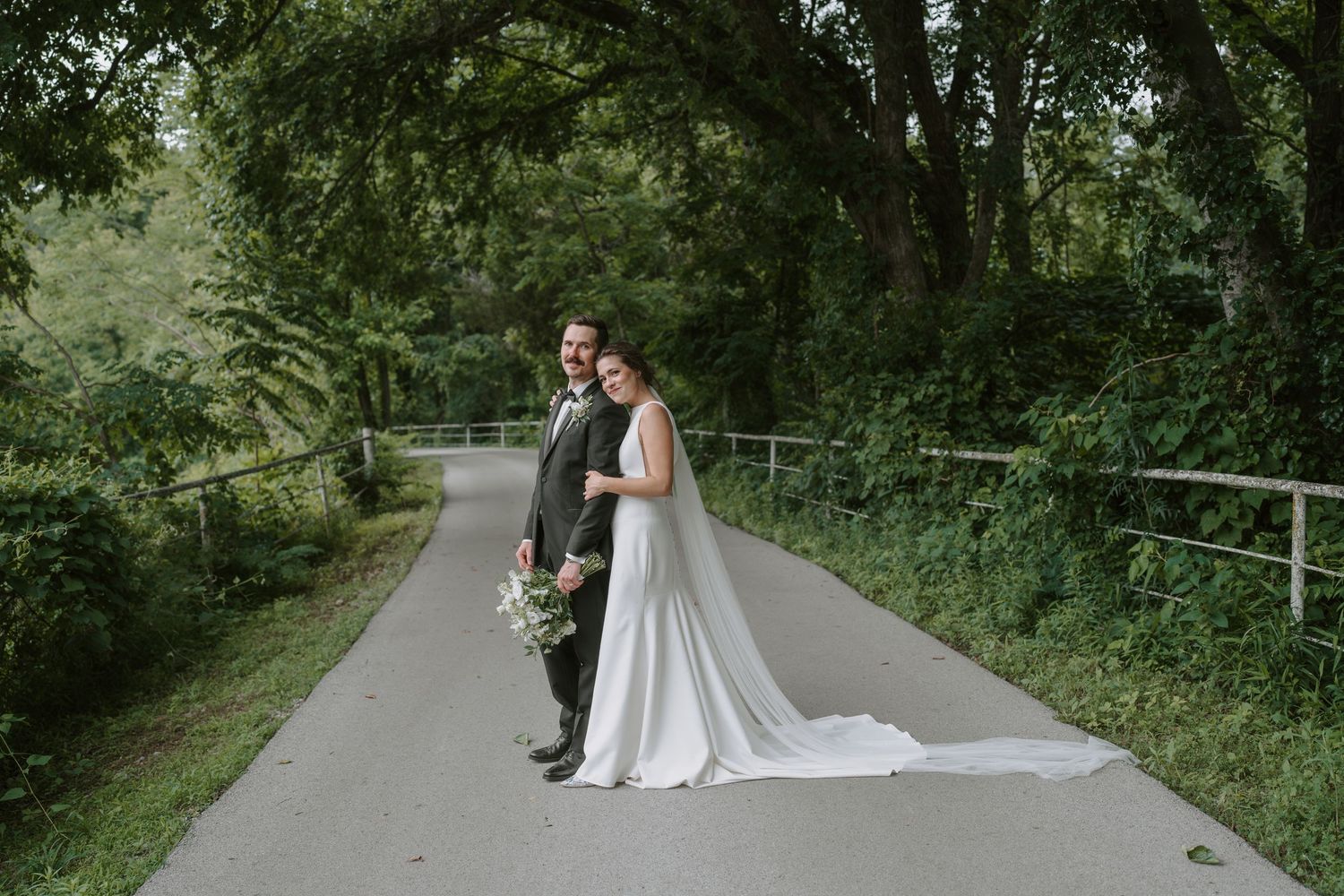Elegant wedding couple poses on a winding path beneath a canopy of green trees.