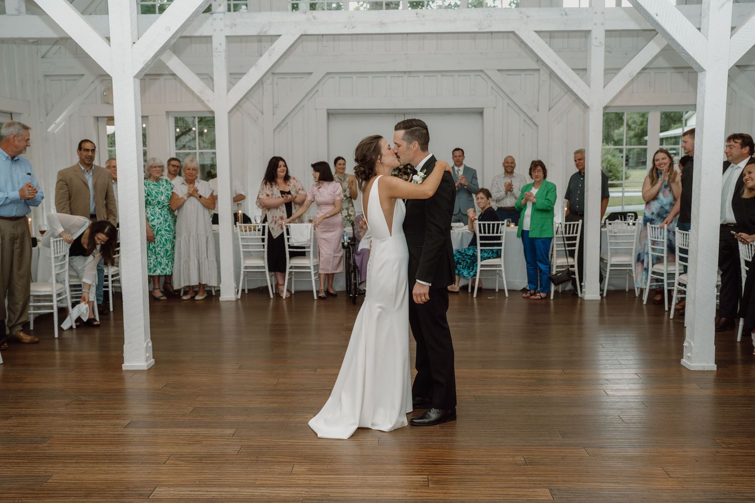 Wedding reception dance in a bright white barn venue with guests seated at round tables.