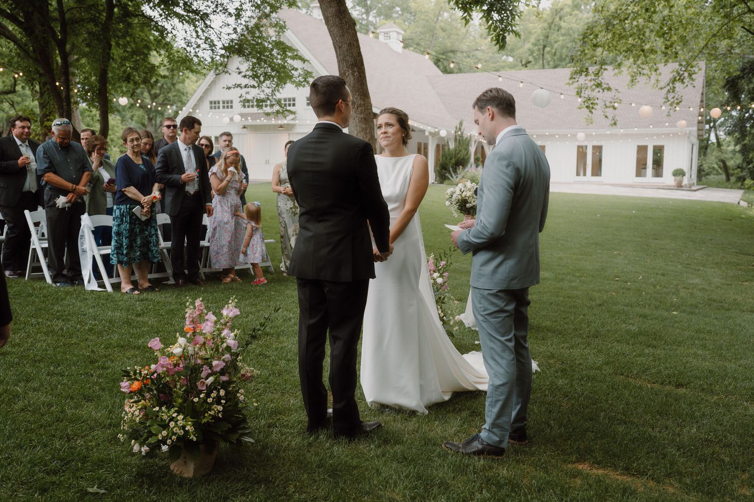 Outdoor wedding ceremony setup with white chairs on a green lawn surrounded by trees.