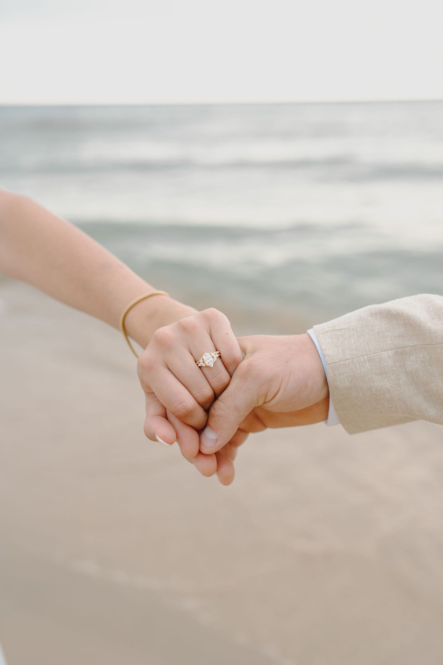 Close-up of intertwined hands with engagement ring against a soft beach background.