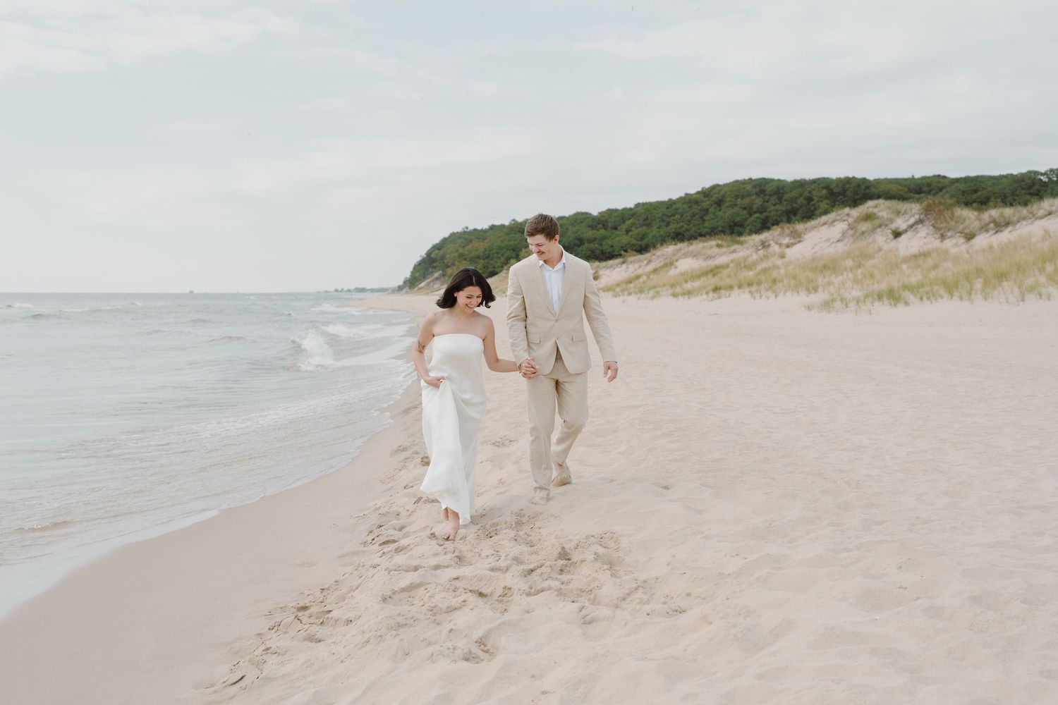 A couple enjoys a romantic walk along the shoreline with coastal dunes in the background.