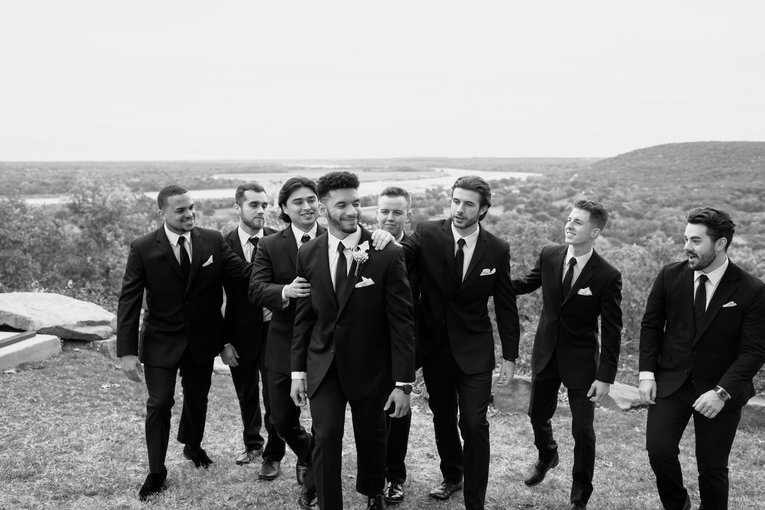 Groomsmen in black suits share laughter and camaraderie during outdoor wedding photos against a scenic backdrop.