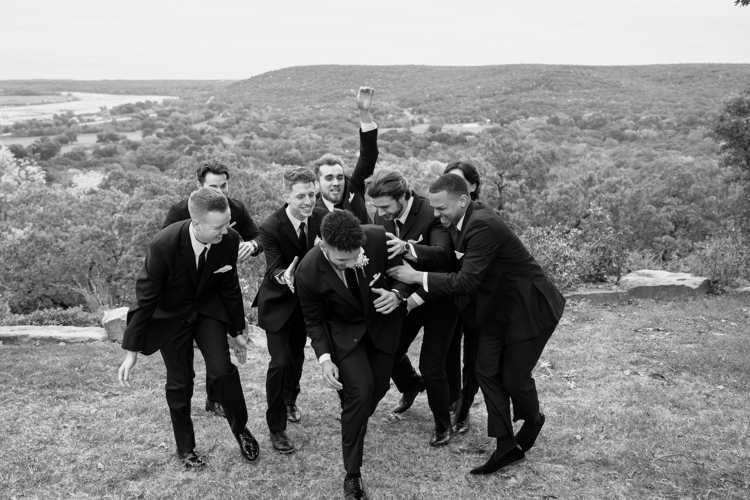 Groomsmen in black suits playfully pose in a sequence of jumping photos on a hilltop landscape.
