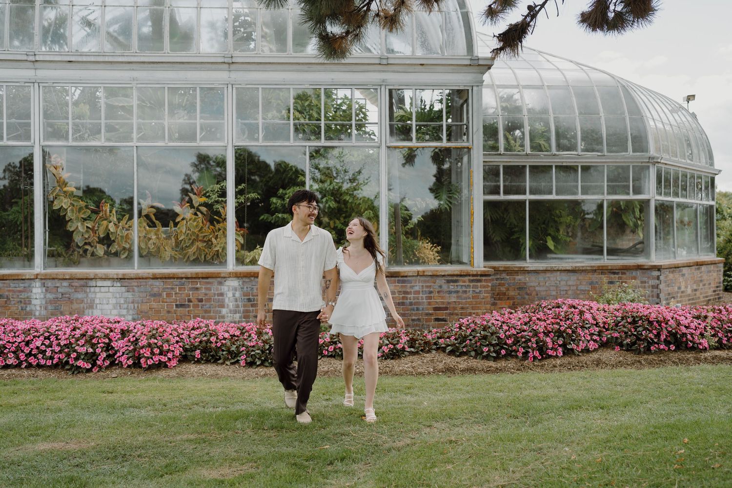 A couple stands together on grass near a greenhouse surrounded by pink flowering plants.