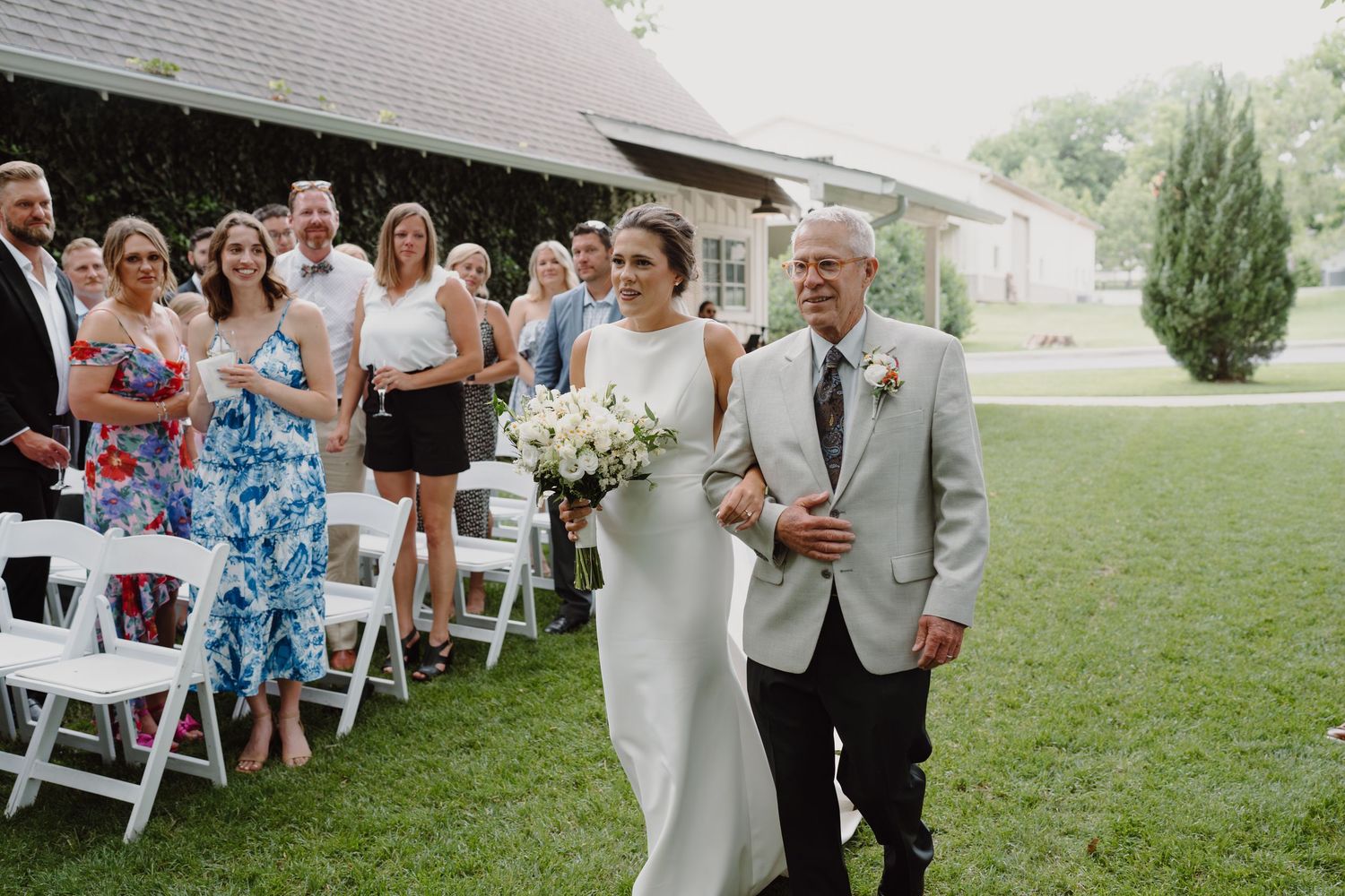 An outdoor wedding ceremony with guests seated in white chairs watching the processional.