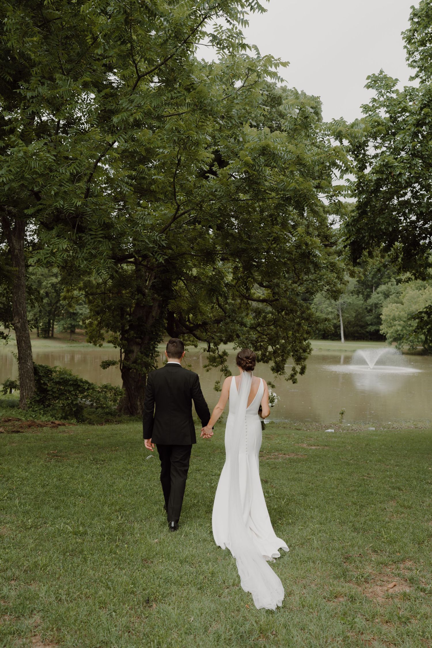 Couple in wedding attire walking near a serene pond surrounded by mature trees.
