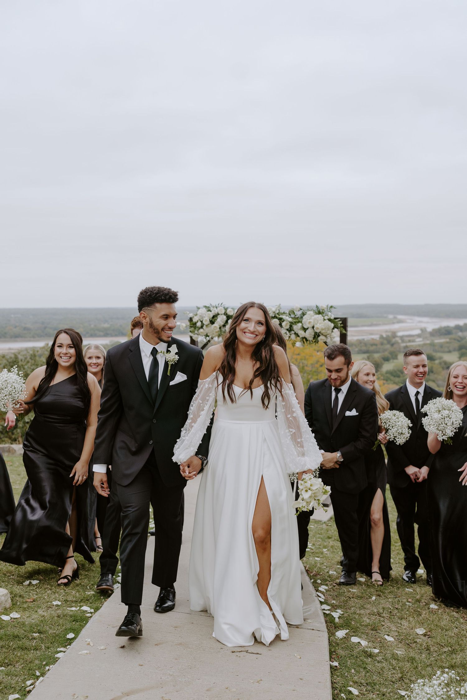 Wedding party in black attire walks along a scenic outdoor path with white floral arrangements.
