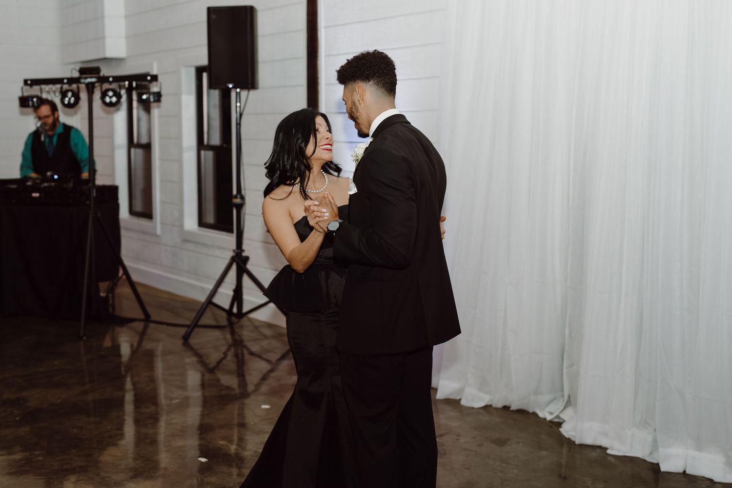 Wedding reception venue with white draping and dark wood floors illuminated by ambient lighting.