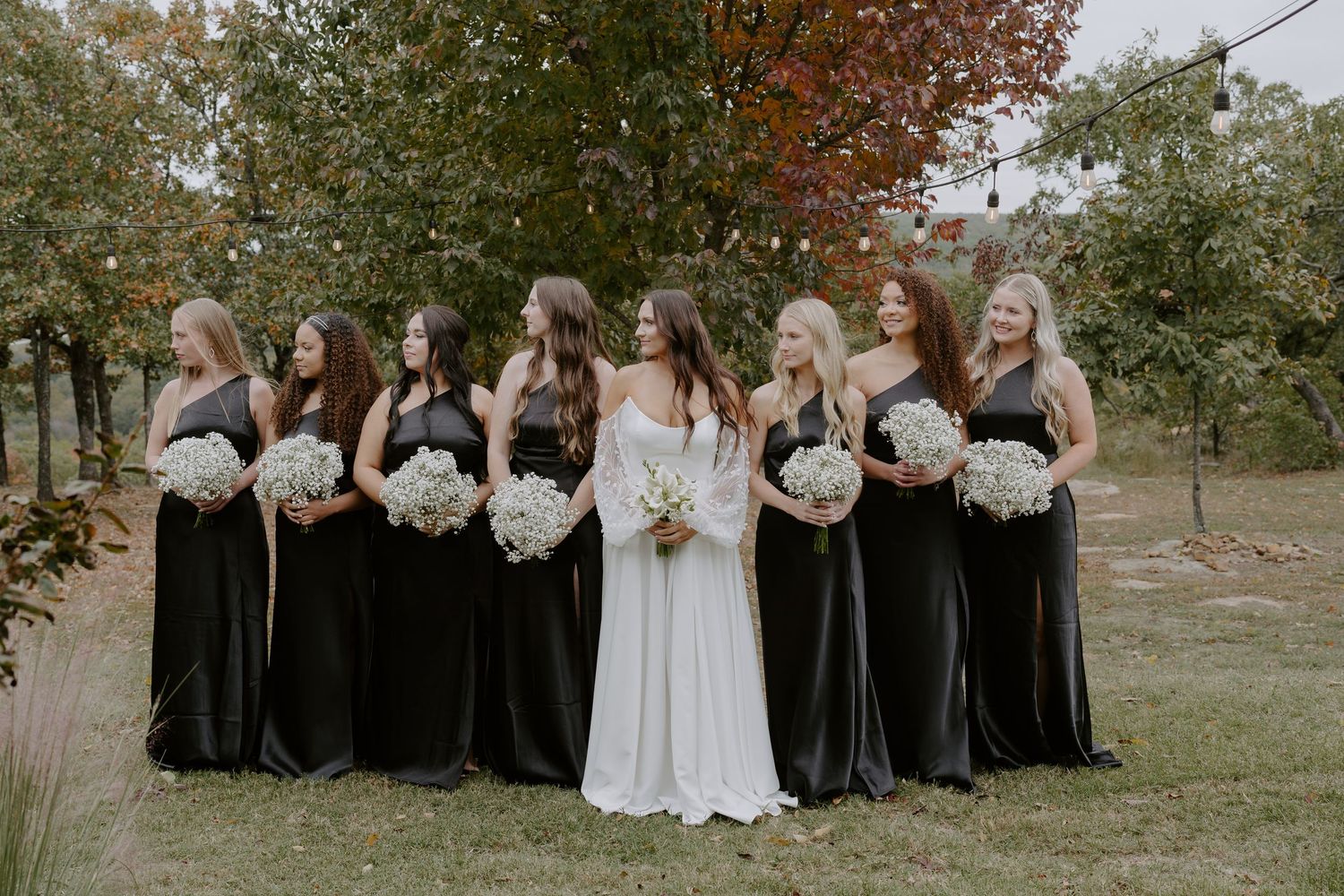 Wedding group stands together on grassy lawn with fall foliage in background.