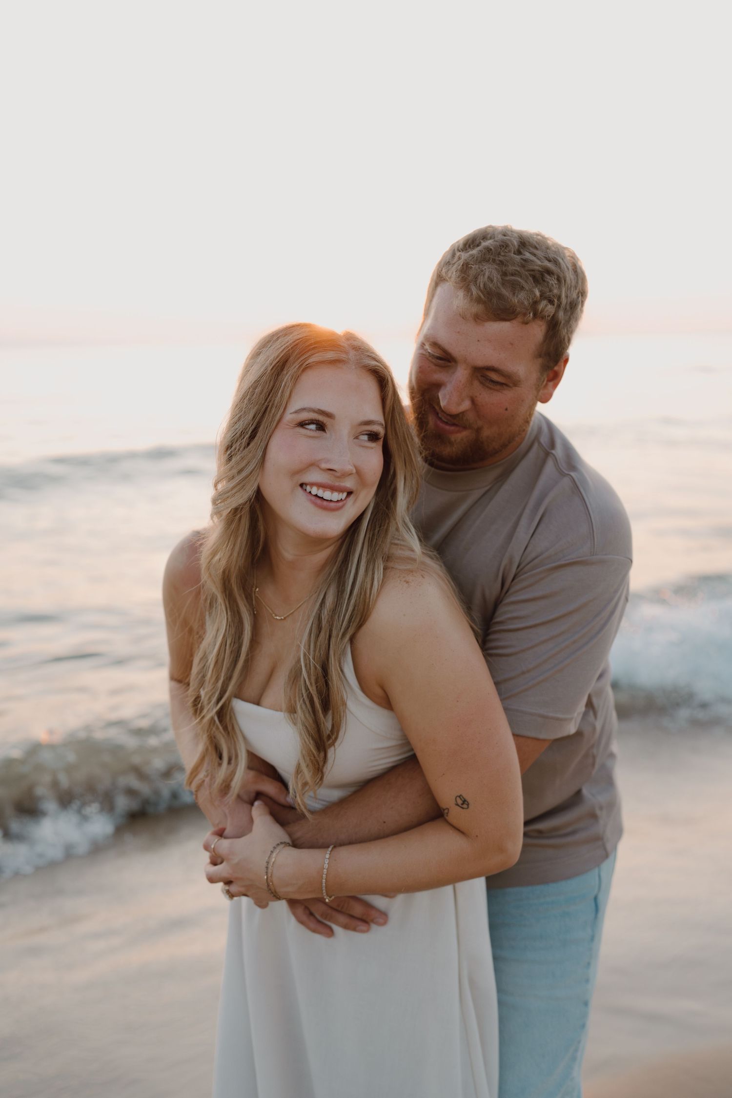 A couple embraces on the beach during golden hour with soft waves in the background.