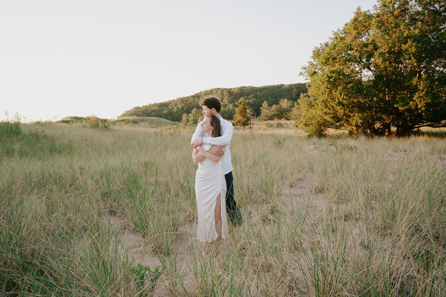Two people walk through a scenic meadow with trees and rolling hills in the background.