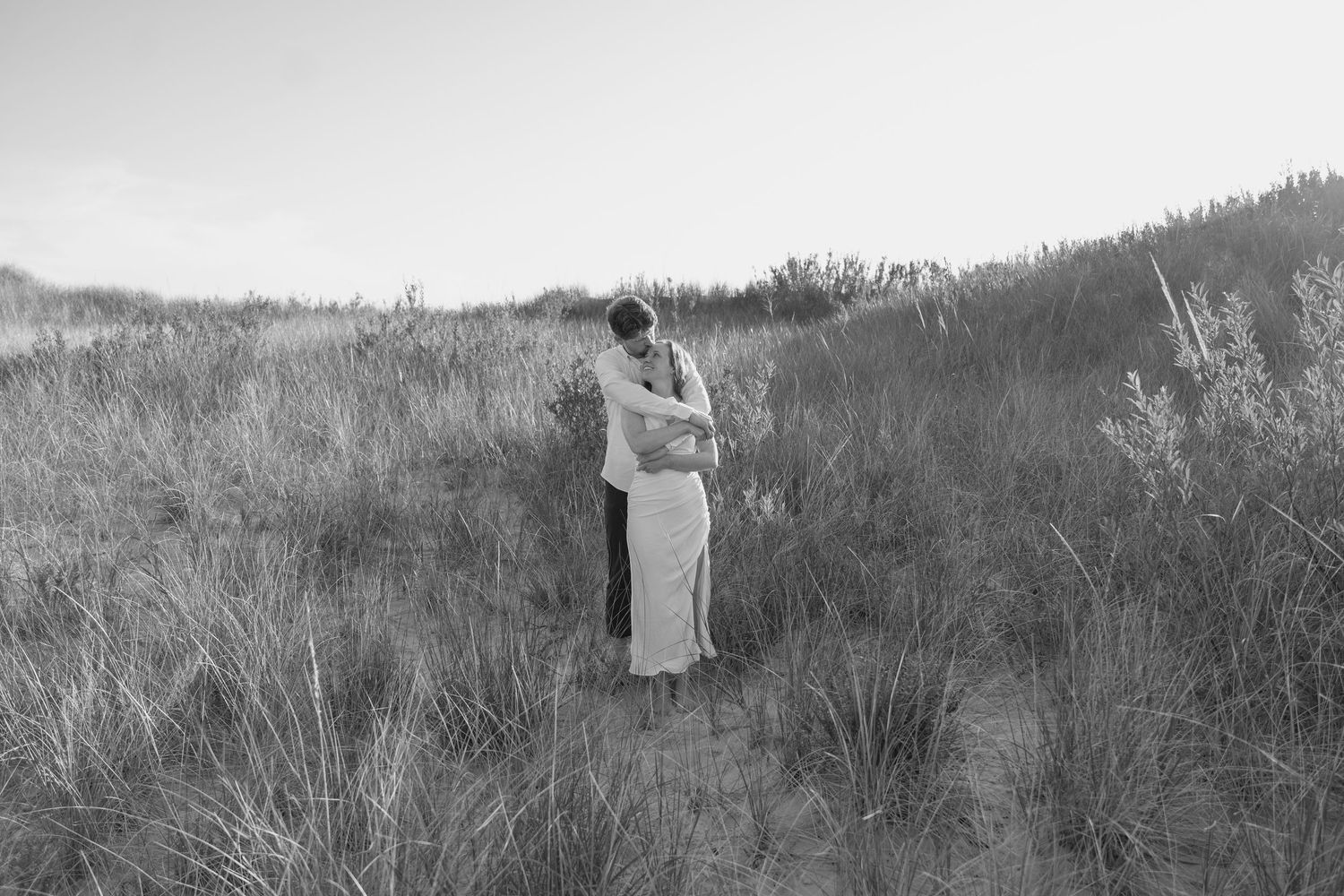 A romantic moment between two people standing among beach grass at dusk.