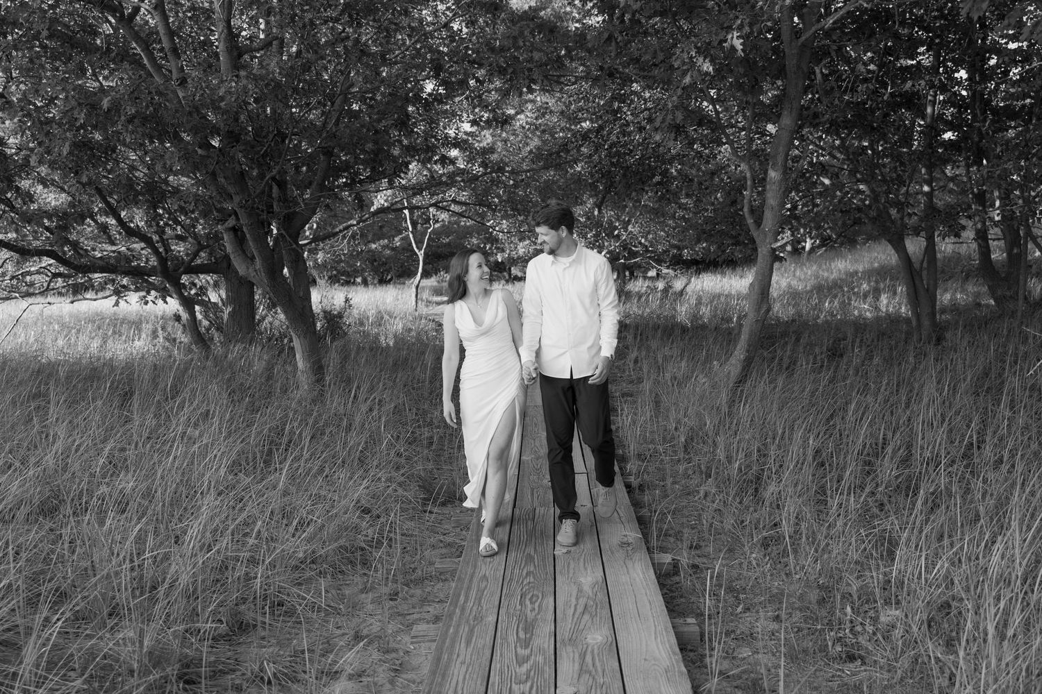 A couple walks together through tall grass in a serene black and white landscape.