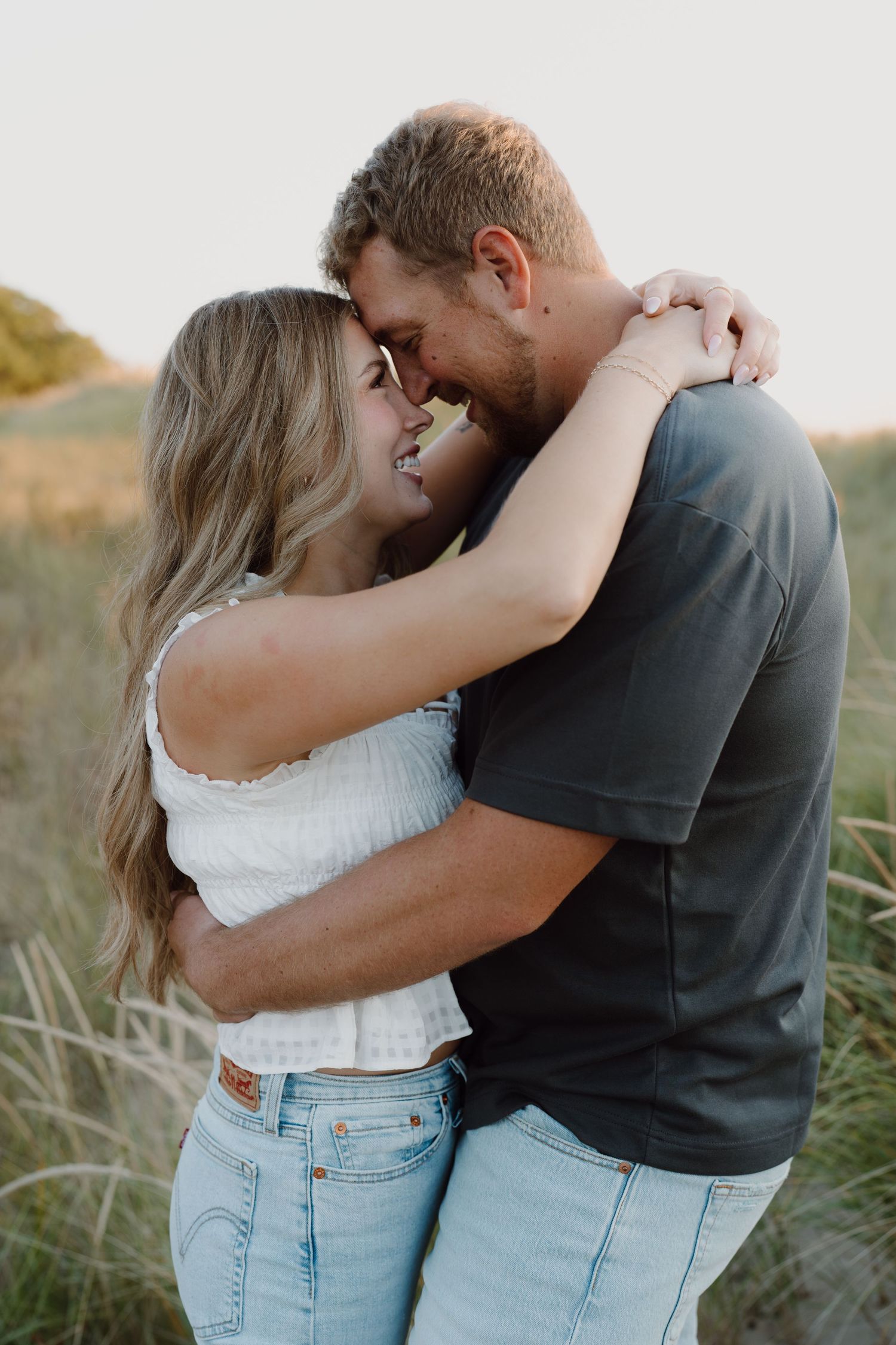 Couple in casual attire embracing lovingly on beach dunes during golden hour.