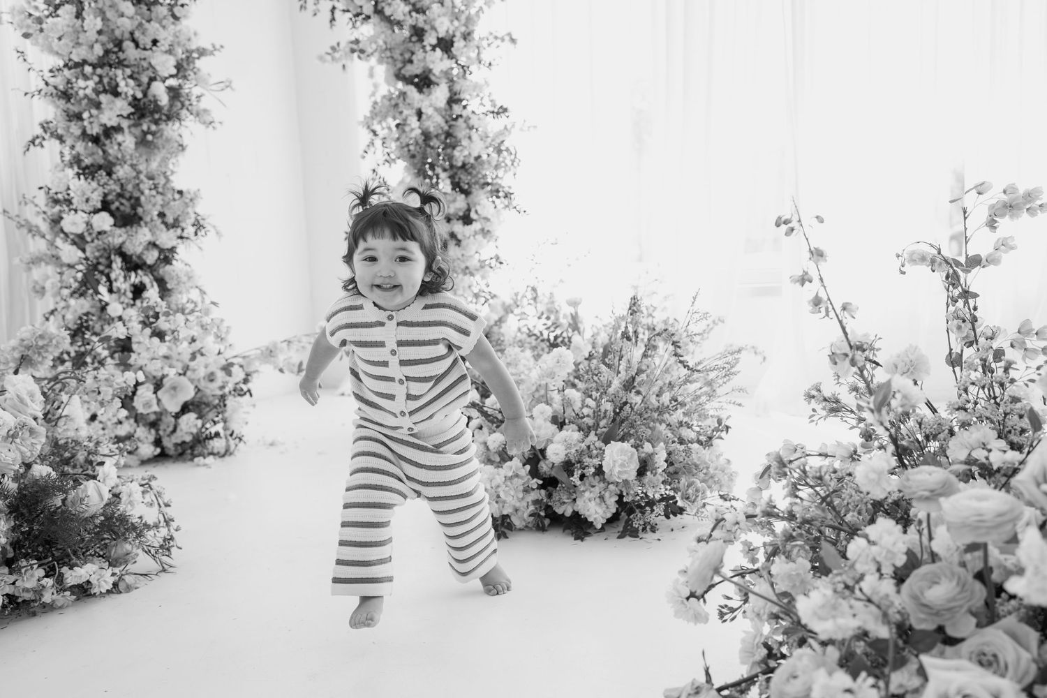Black and white portrait of child in striped outfit among floral arrangements.