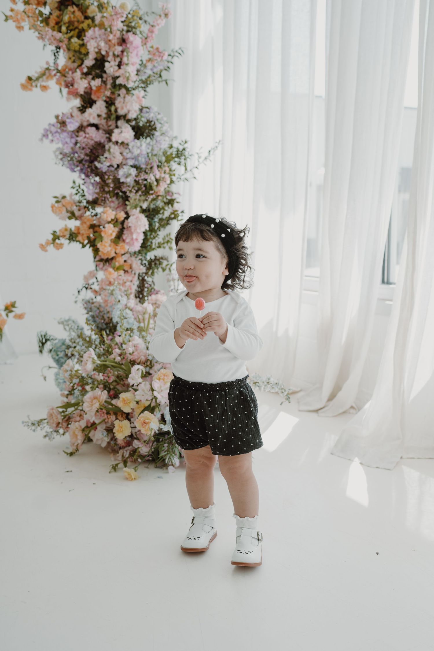 Toddler in white shirt and polka dot shorts standing near floral arrangements.